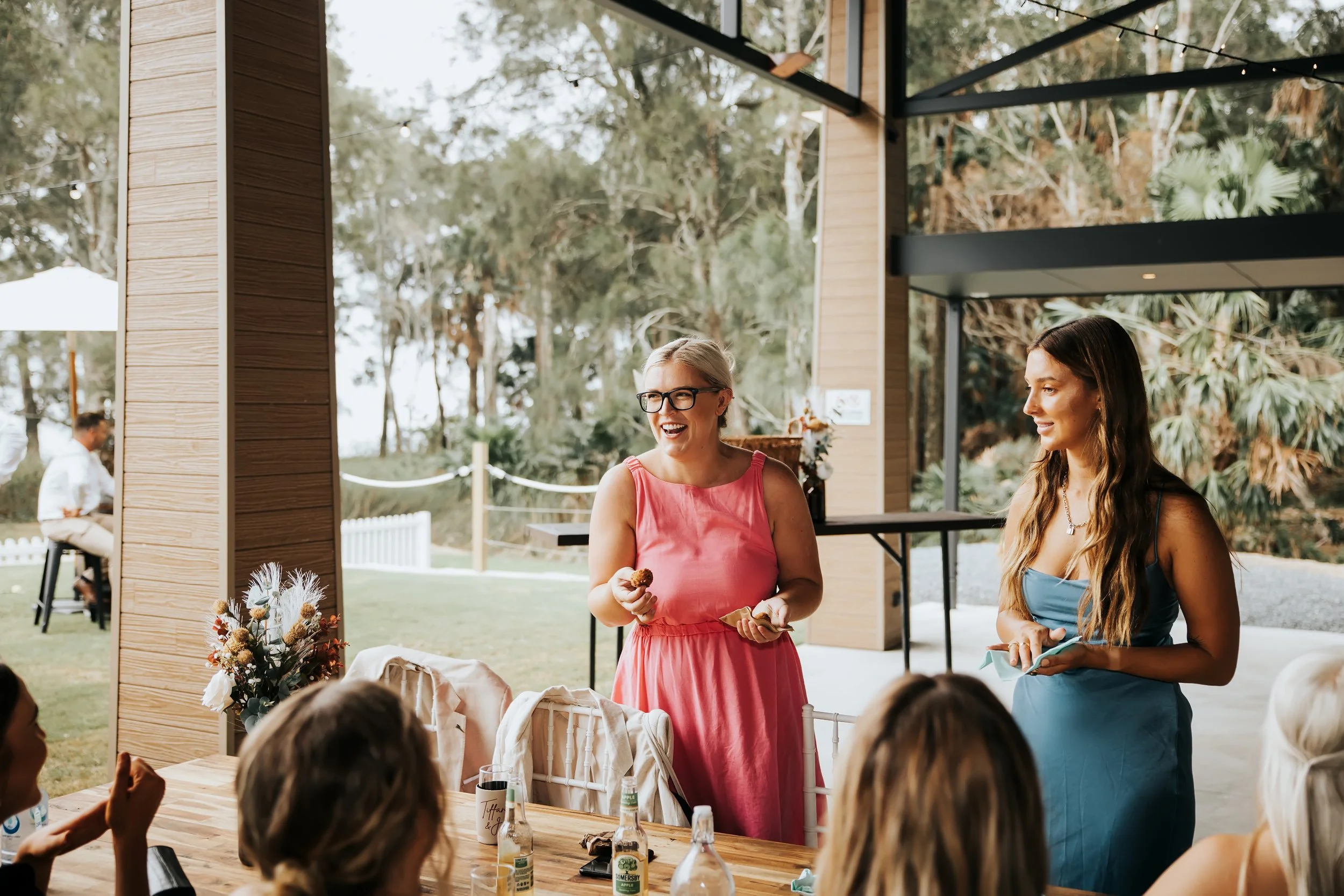 Two women in colorful dresses, one in pink and the other in blue, are standing and smiling at a gathering outdoors under a pavilion with trees in the background. Several guests are seated, and a floral arrangement is visible on the table.