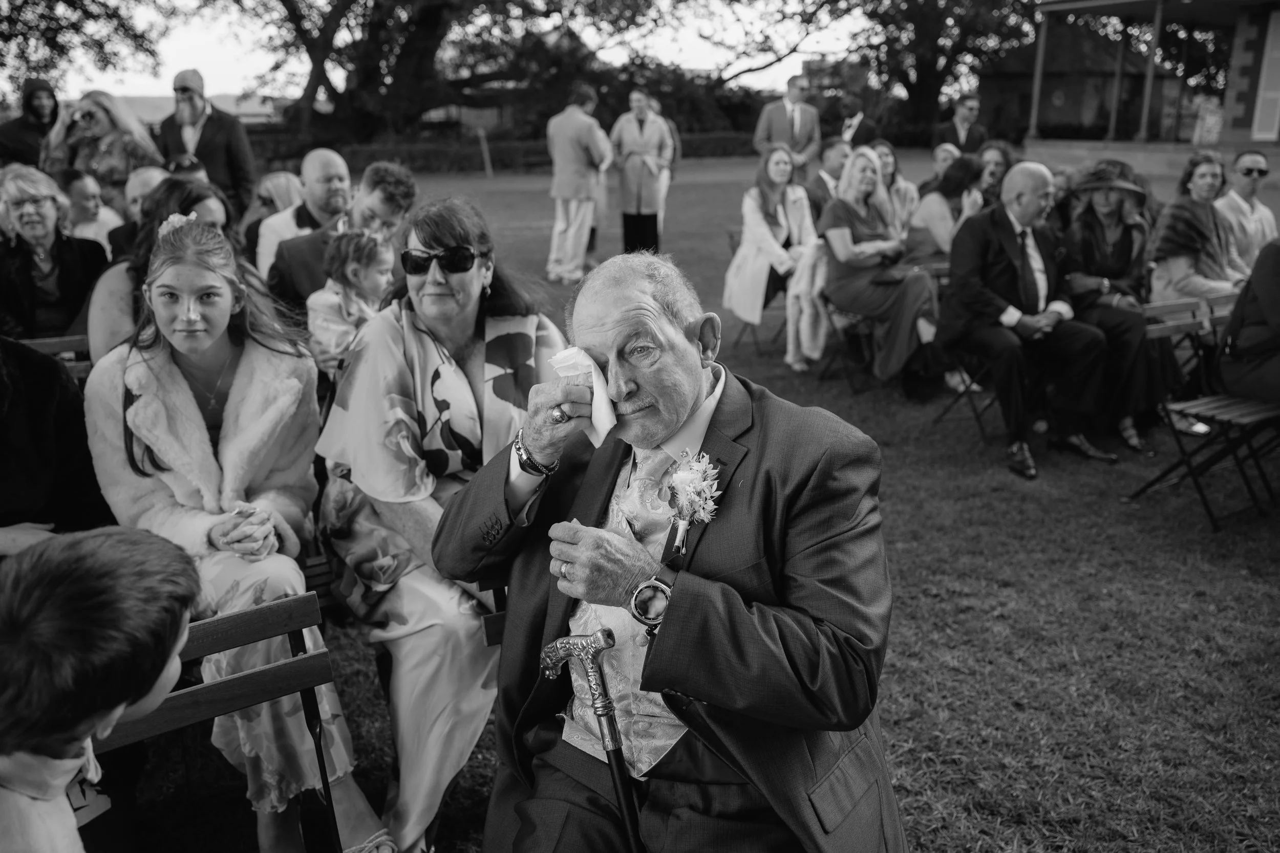 A black and white photo of an outdoor gathering with people seated on chairs. In the foreground, an elderly man in a suit is wiping tears from his eyes with a tissue, holding a walking cane. Behind him are women and children, with some women wearing 