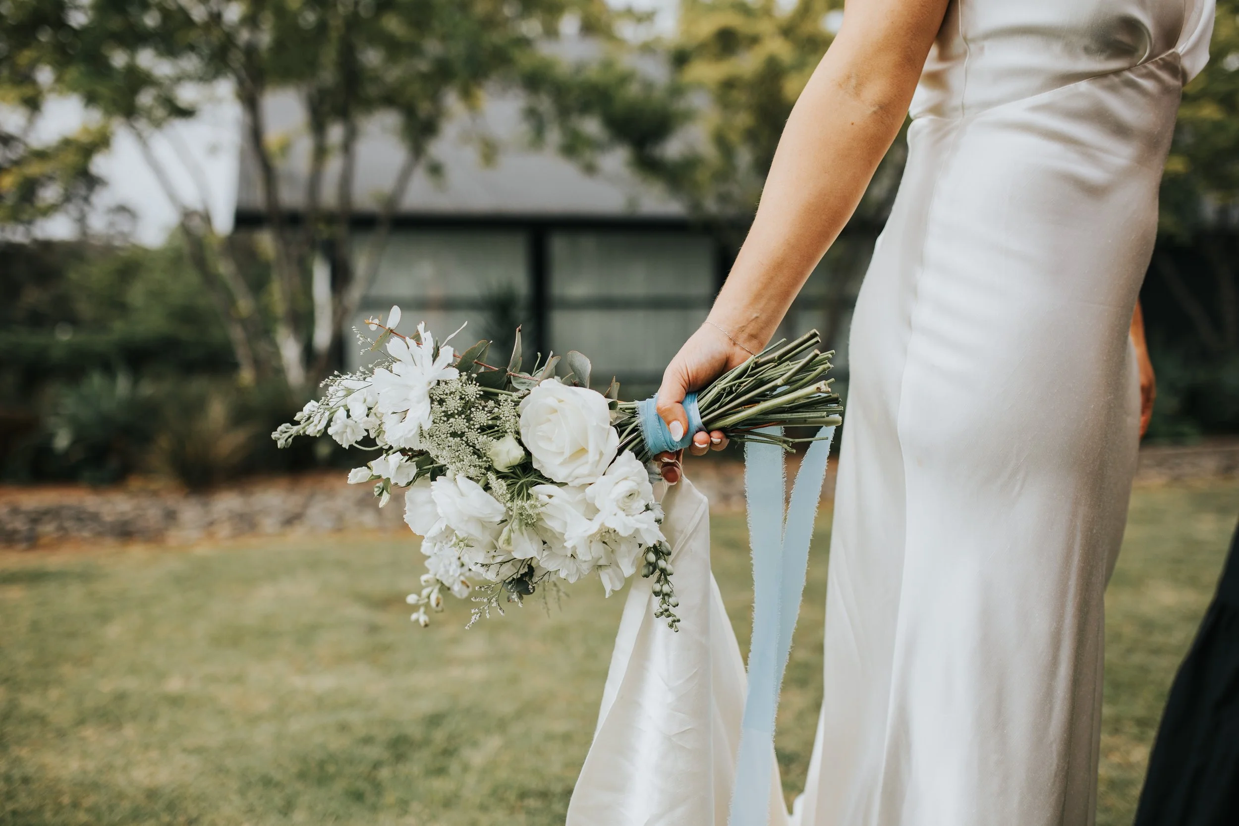 A bride in a white satin wedding dress holding a bouquet of white flowers and greenery outdoors on a lawn with trees in the background.