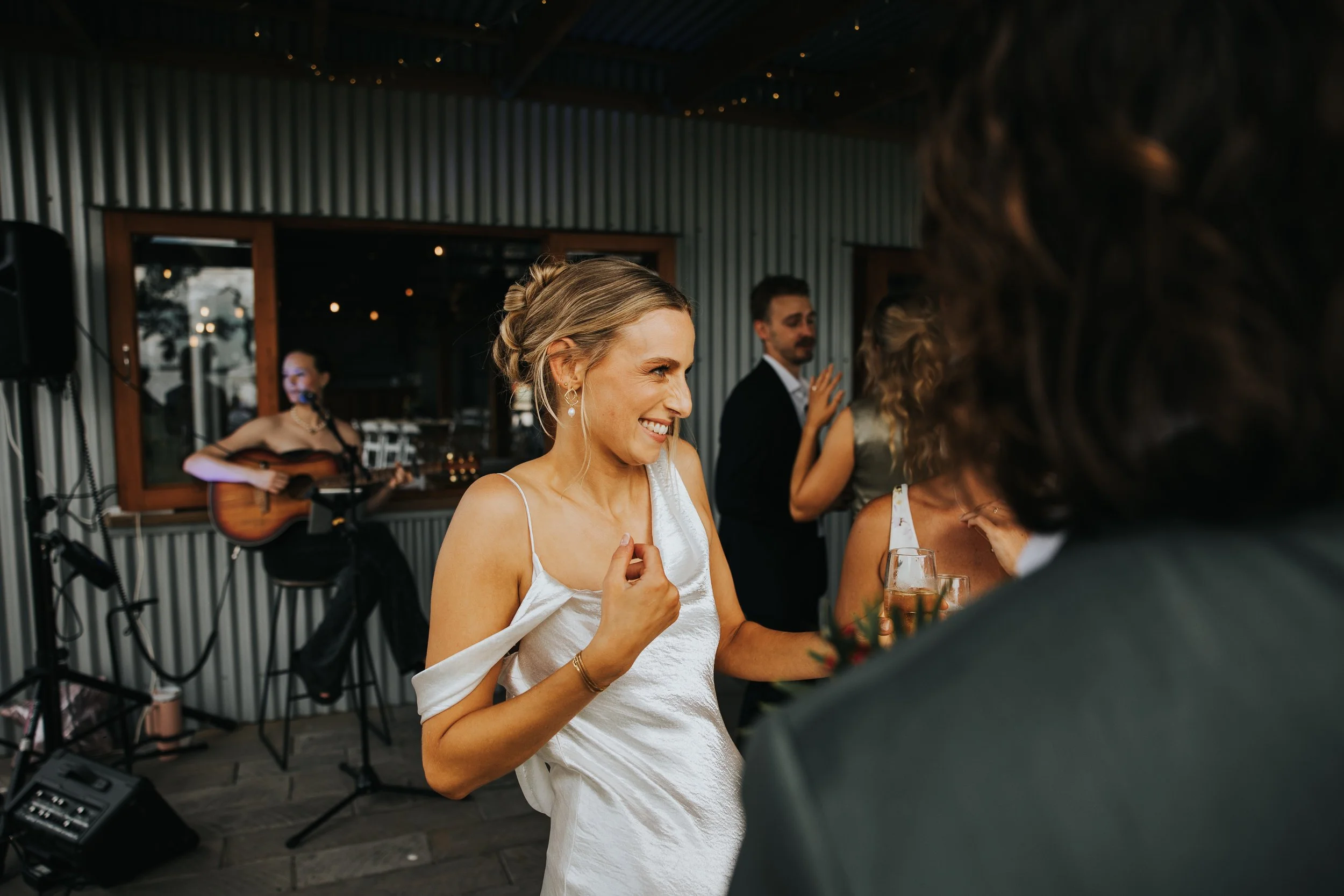 A woman in a white dress dancing and smiling at a social gathering, with a musician playing guitar in the background and other guests conversing.