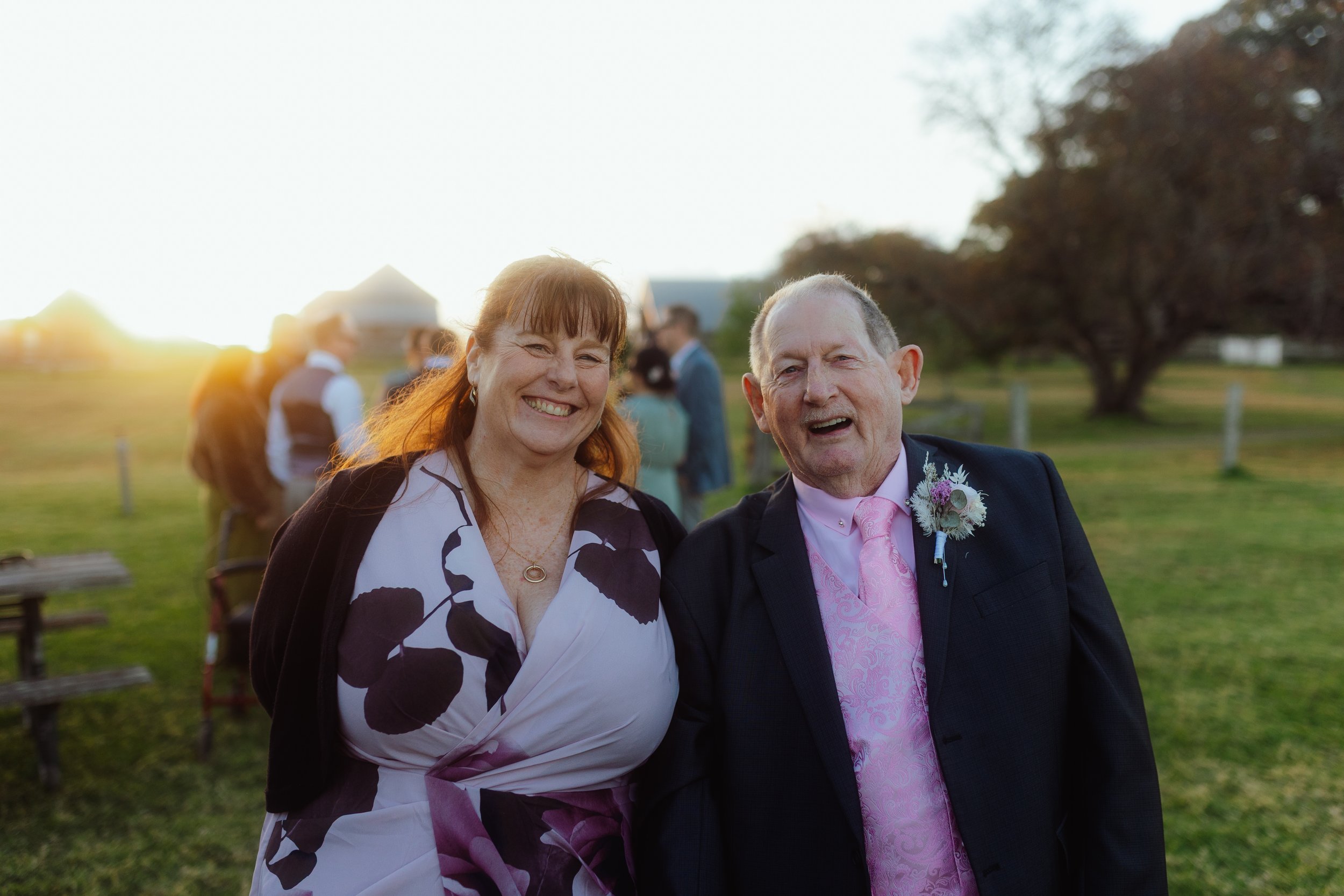 A smiling woman with red hair and a patterned dress standing next to an older man in a dark suit and pink tie with a boutonniere, outdoors during sunset, with other people and trees in the background.
