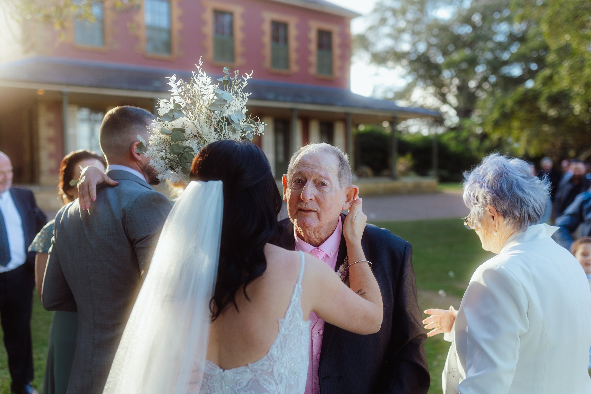 People hugging and smiling at an outdoor wedding reception with a brick building and trees in the background.