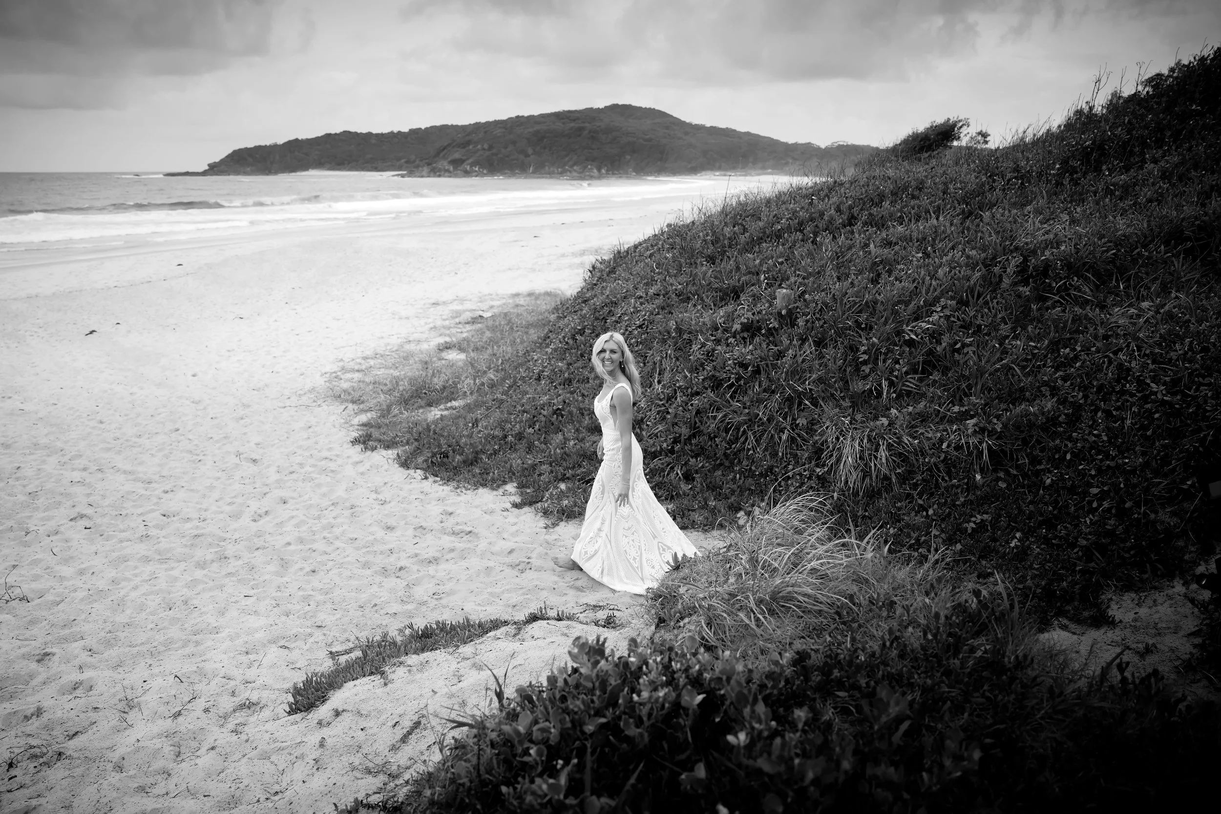 A woman in a white dress standing on a sandy beach near bushes with a hill and ocean in the background, black and white photo.