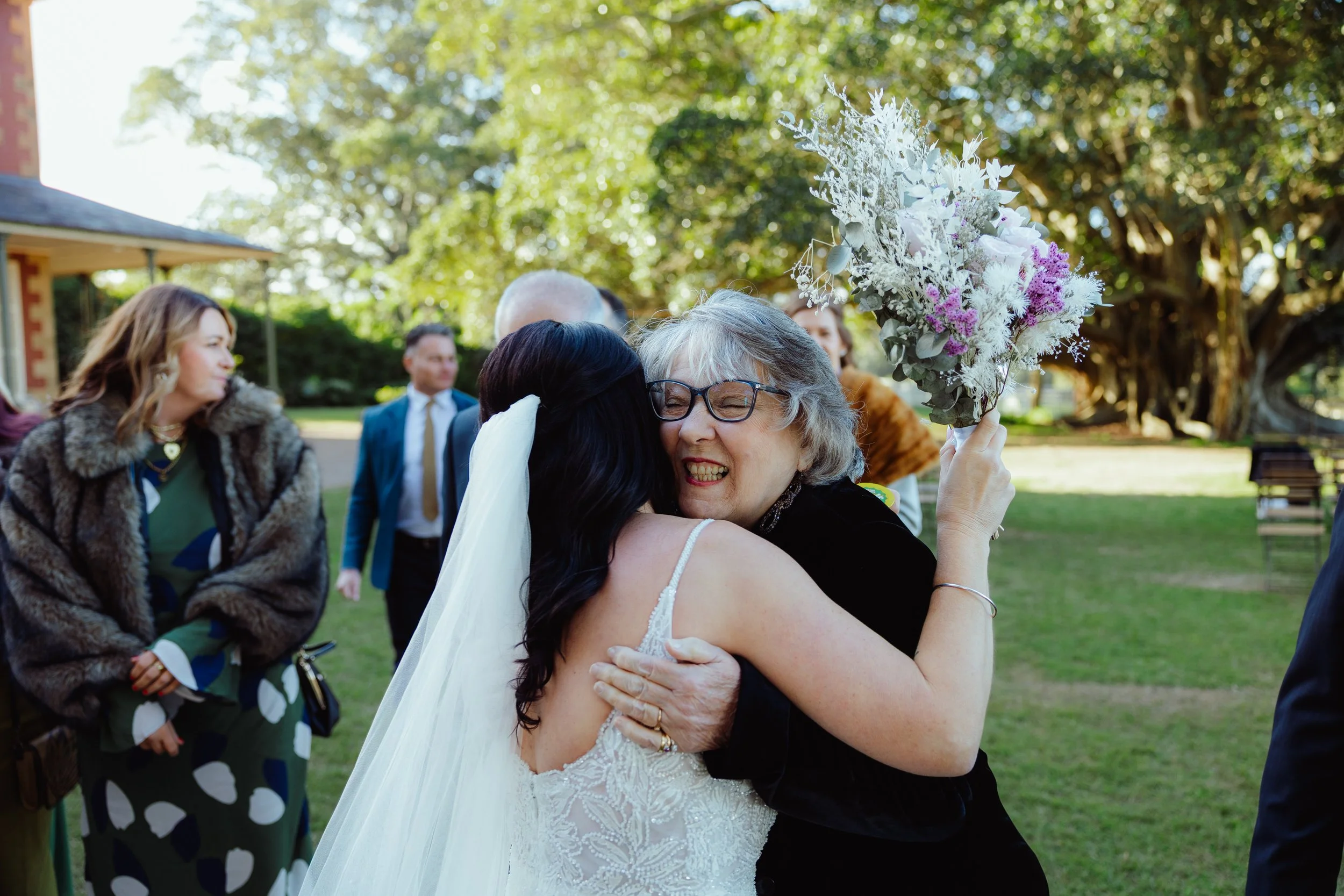 A woman in a white wedding dress hugging an older woman with glasses who is holding a bouquet of white and purple flowers, in an outdoor garden setting during a wedding celebration.