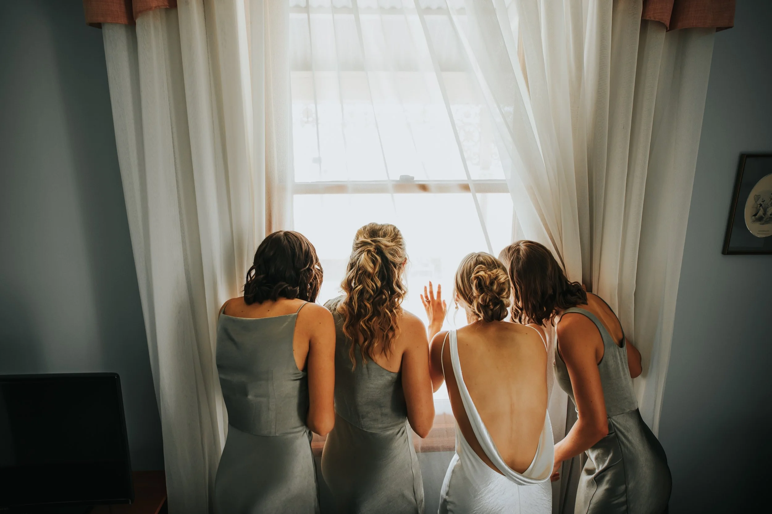 Six women in matching gray and white dresses, facing a window with sheer curtains, looking outside and waving.