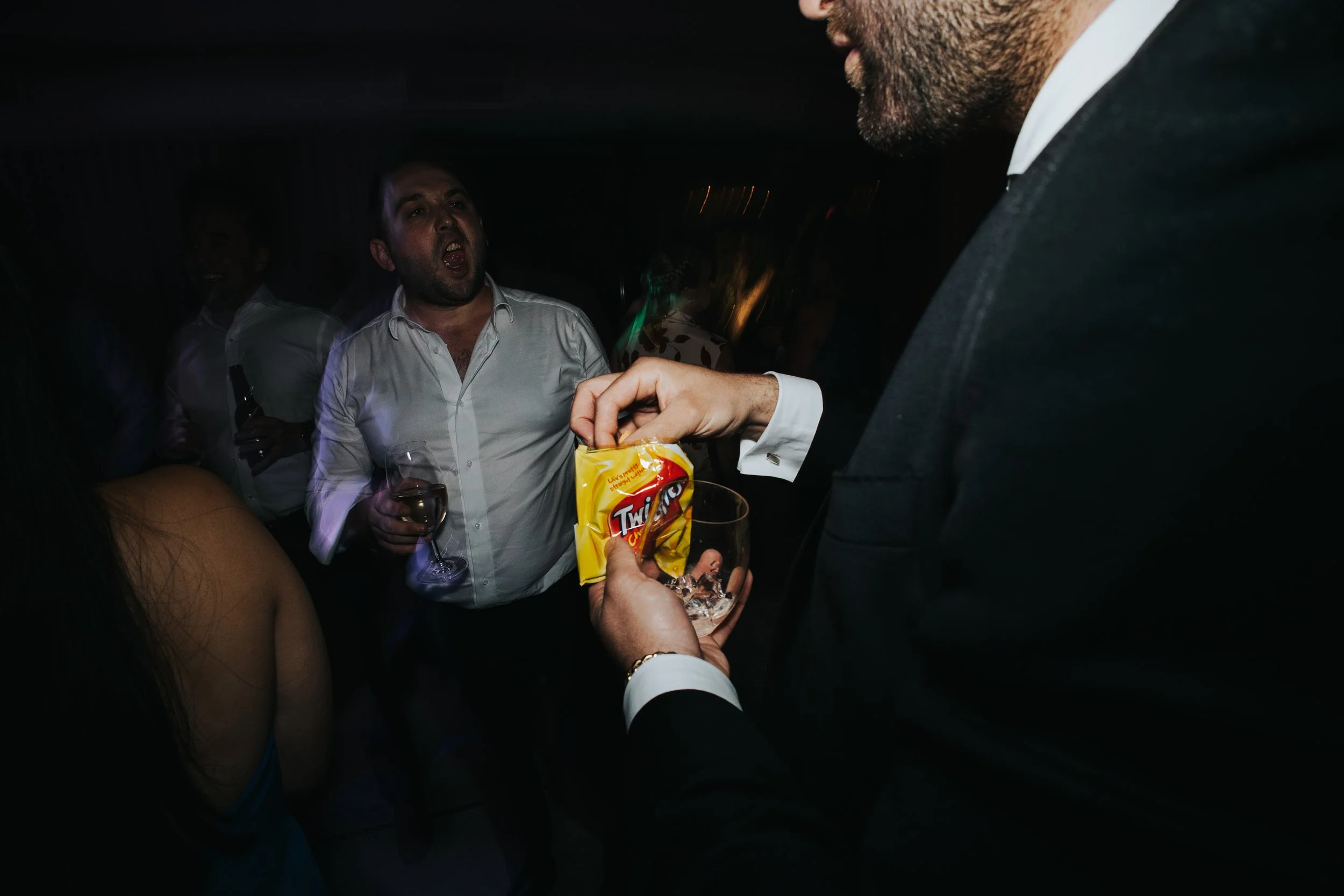 A man in a gray shirt and a woman with dark hair are at a party, with others around them. The woman is holding a glass of wine, and another person's hand is offering a yellow snack bag and a glass filled with ice.