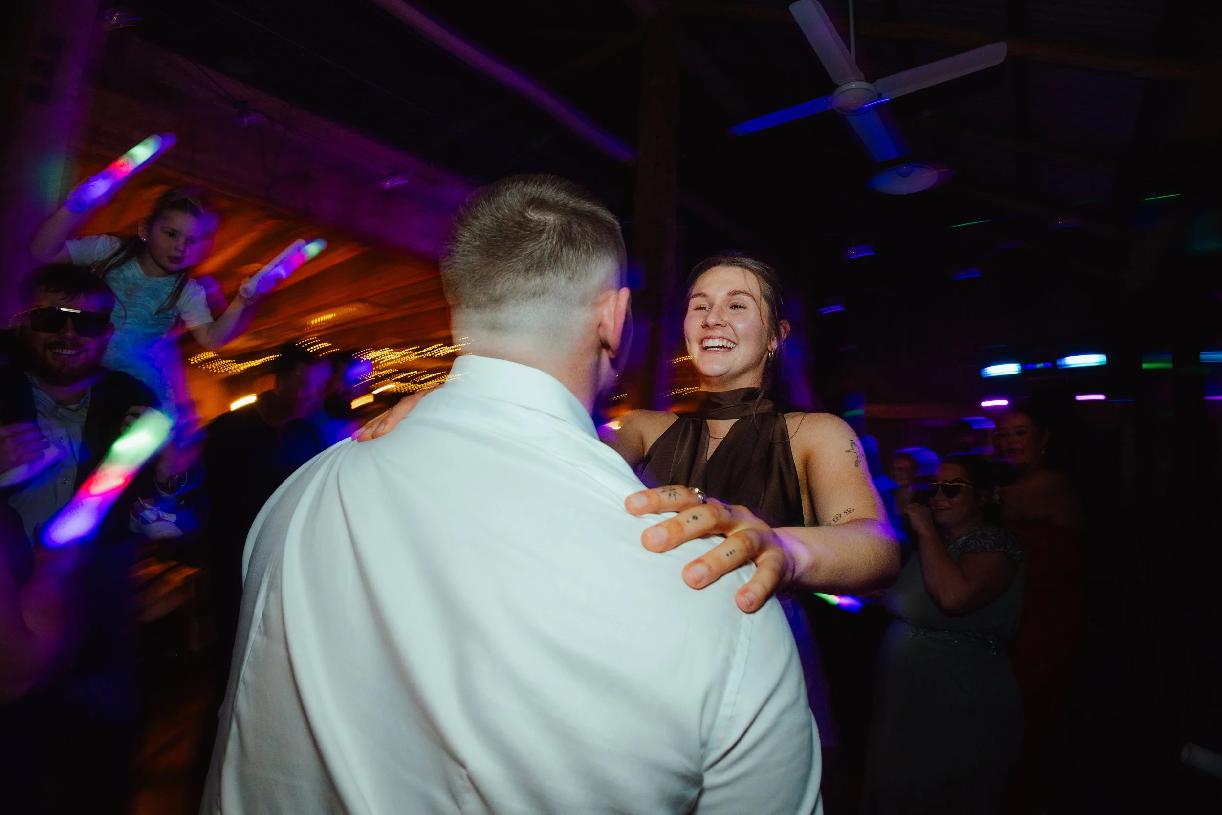 A man and a woman dancing together at a party or celebration, surrounded by colorful lights and other people, including a girl with light-colored hair wearing gloves, in a lively indoor setting.