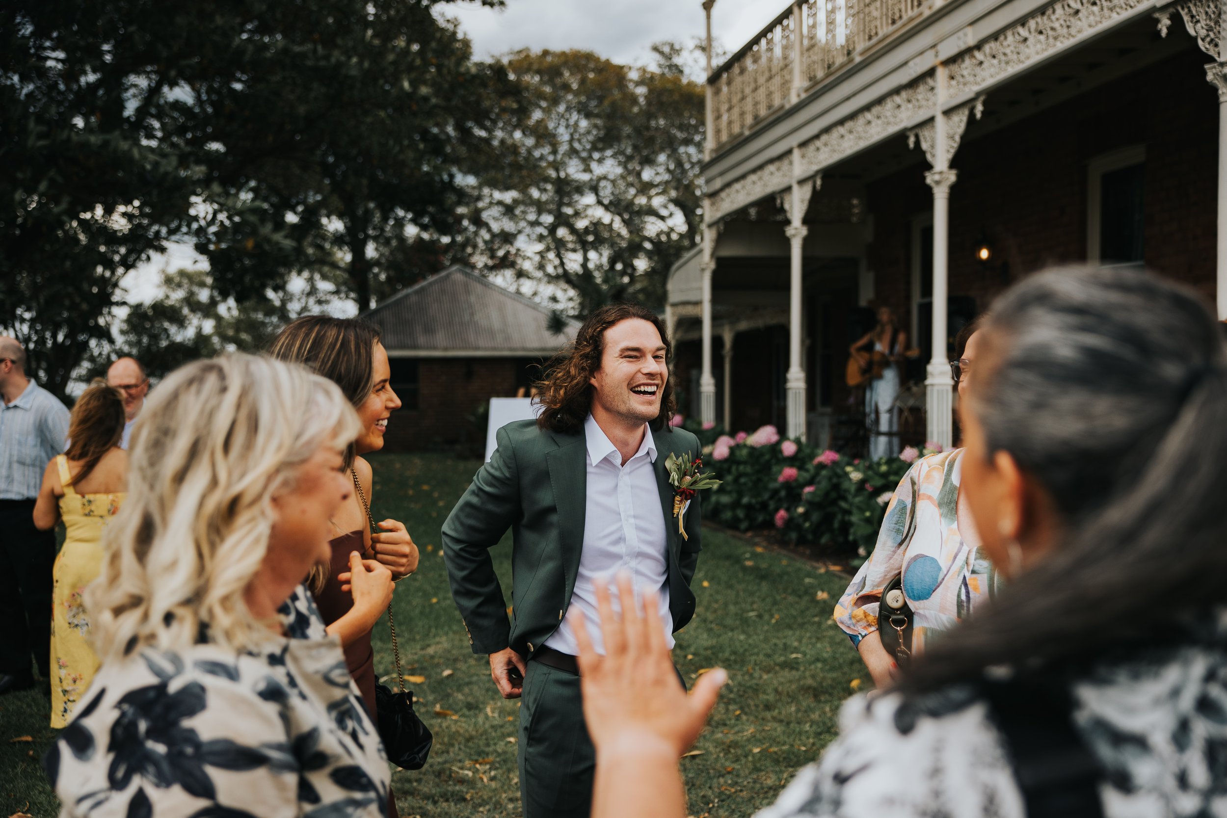 A group of people at an outdoor gathering, with a man in a suit smiling and engaging in conversation, surrounded by women and others, in front of a house with a porch and trees in the background.