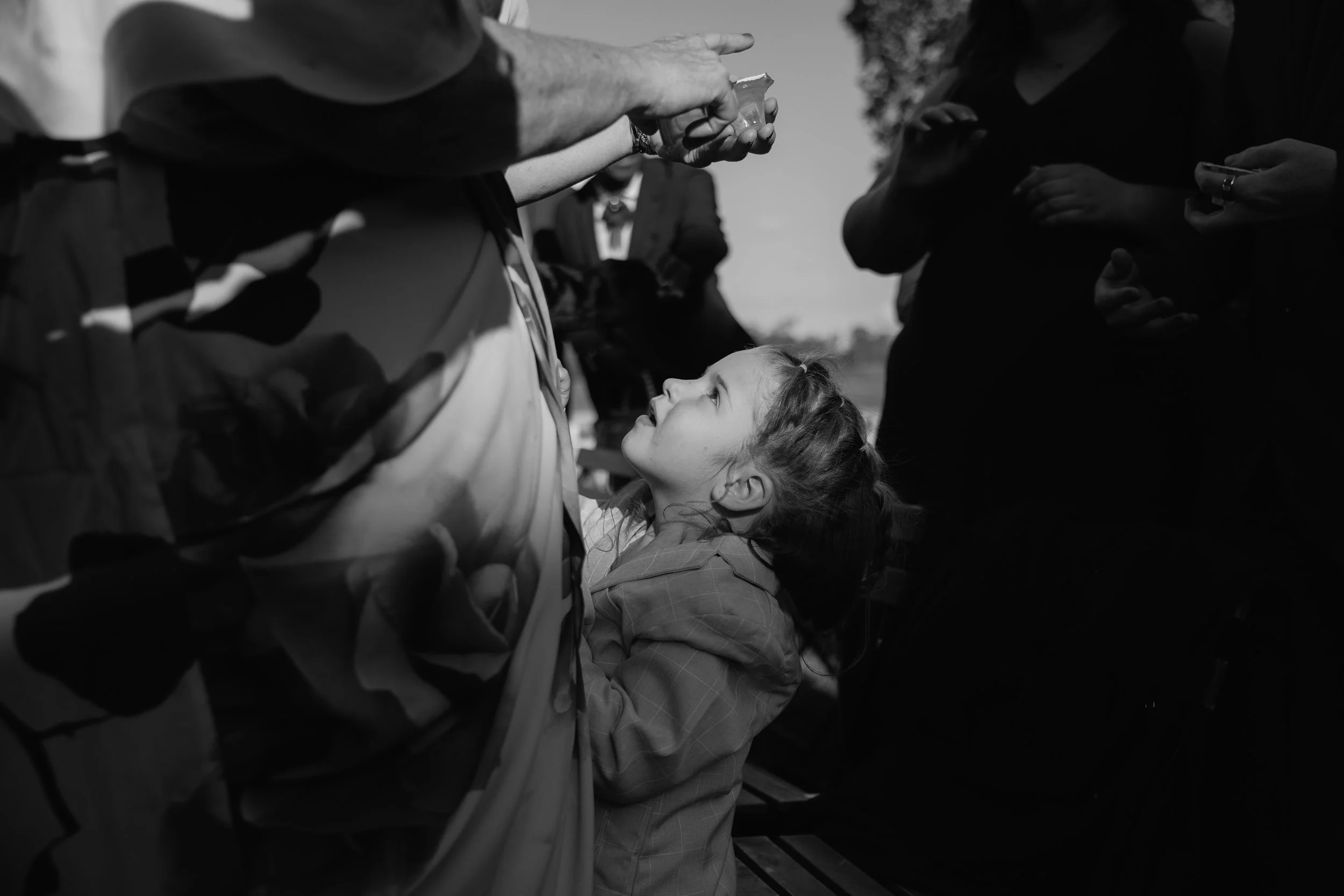 A young girl is looking up at an adult's hand as it offers her a small object. Other people surround them, some with crossed arms or holding phones, and there is a blurred person in the background. The photo is in black and white.