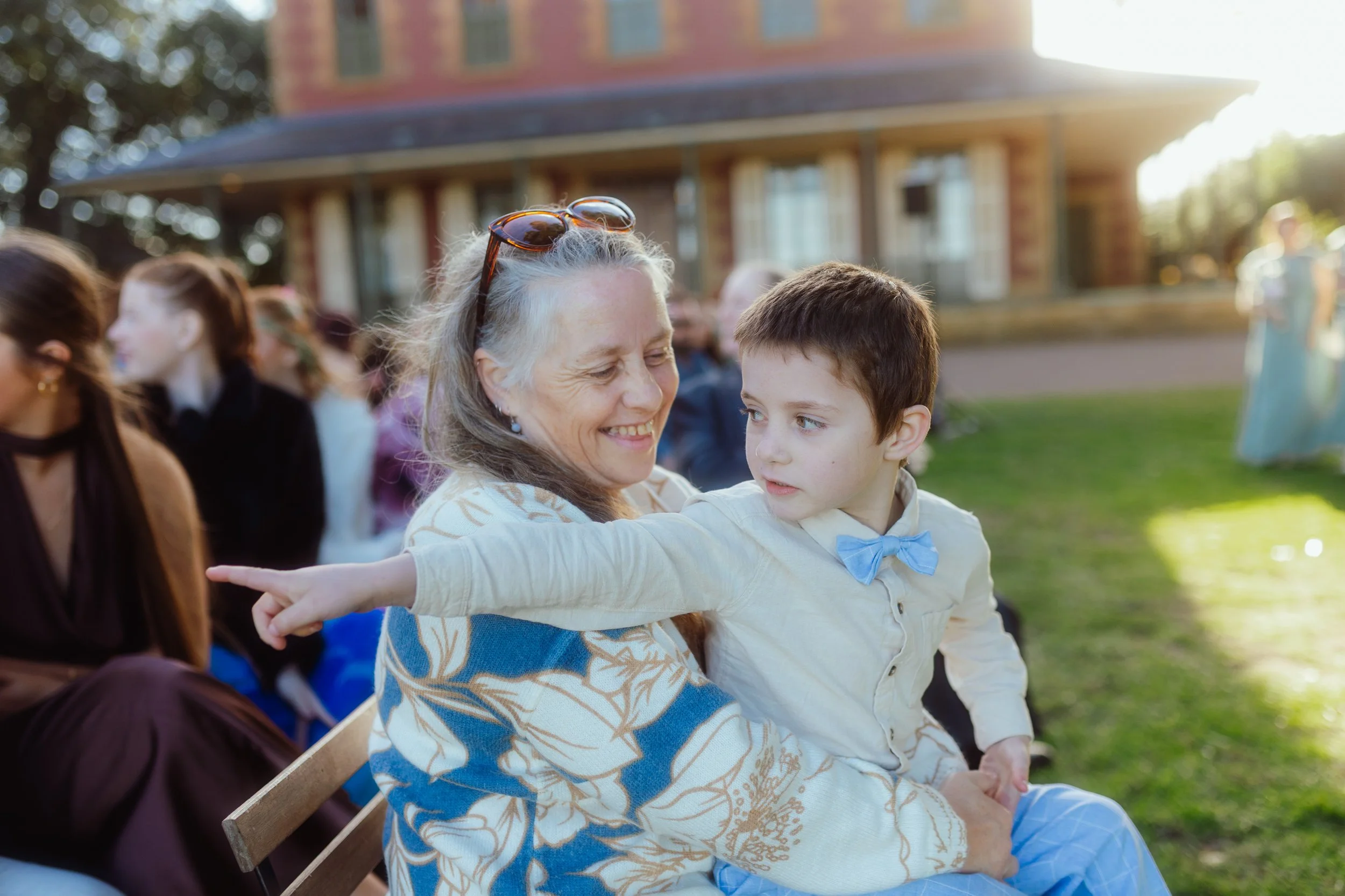 A woman with sunglasses on her head smiling and holding a young boy in an outdoor setting, with other people seated nearby and a building in the background.