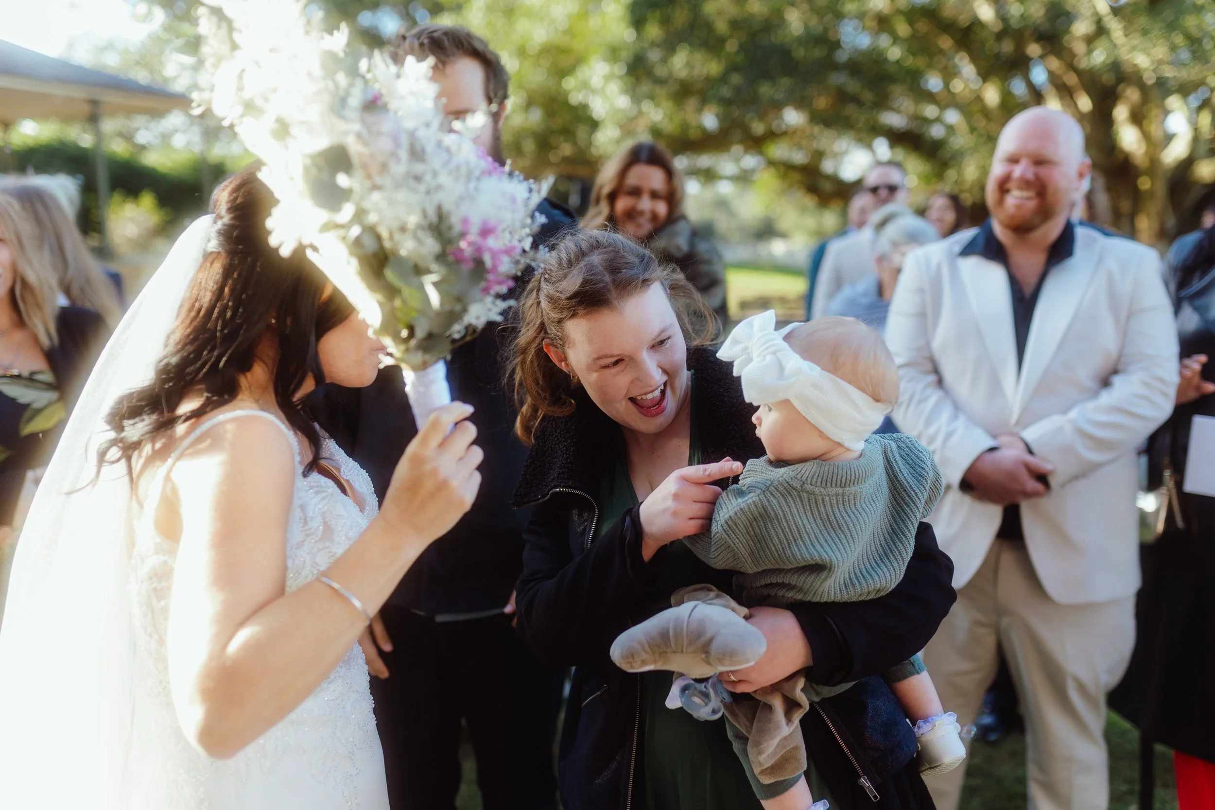 A group of people celebrating at an outdoor wedding reception, with a woman in a white wedding dress and veil holding a large bouquet of white and pink flowers, surrounded by friends and family smiling and enjoying the moment.