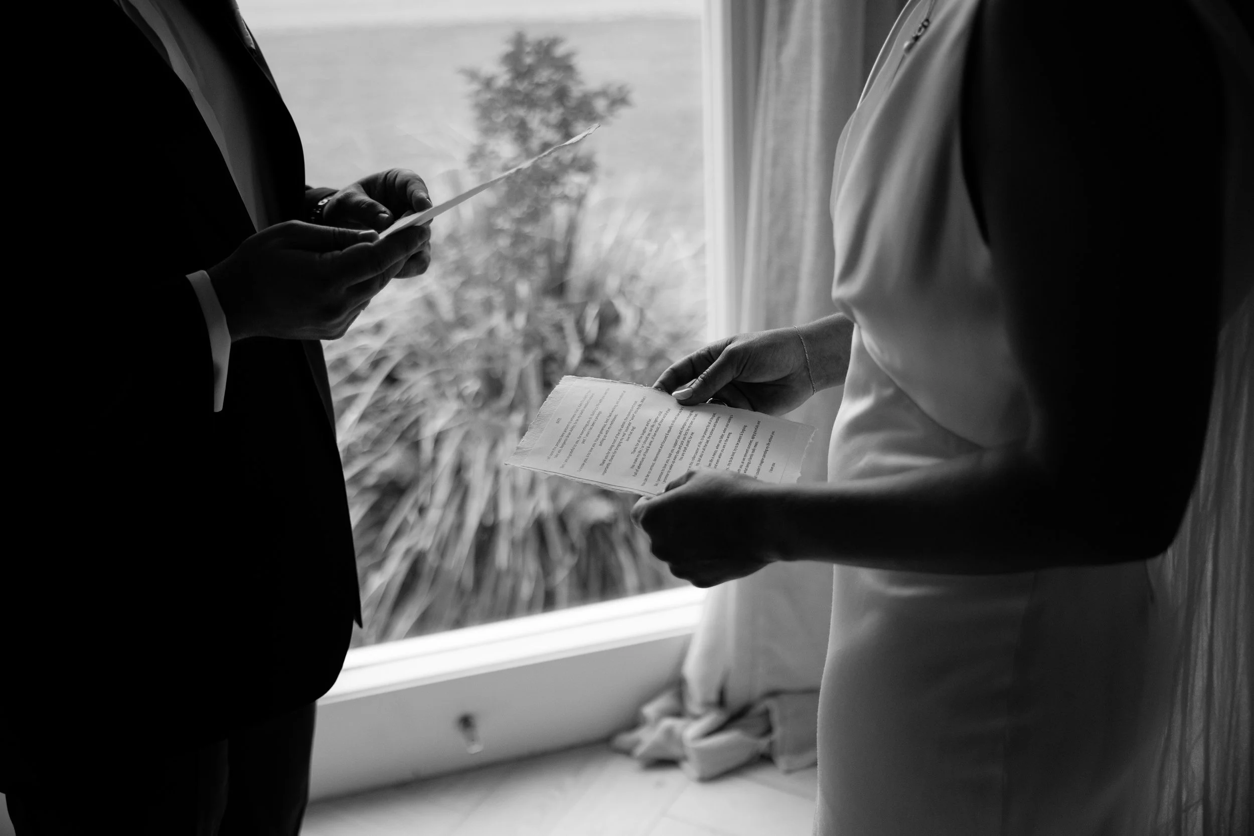 A black and white photo showing a couple at a wedding, with the groom holding a piece of paper and the bride holding a booklet, both standing near a window with a blurred outdoor scene visible in the background.
