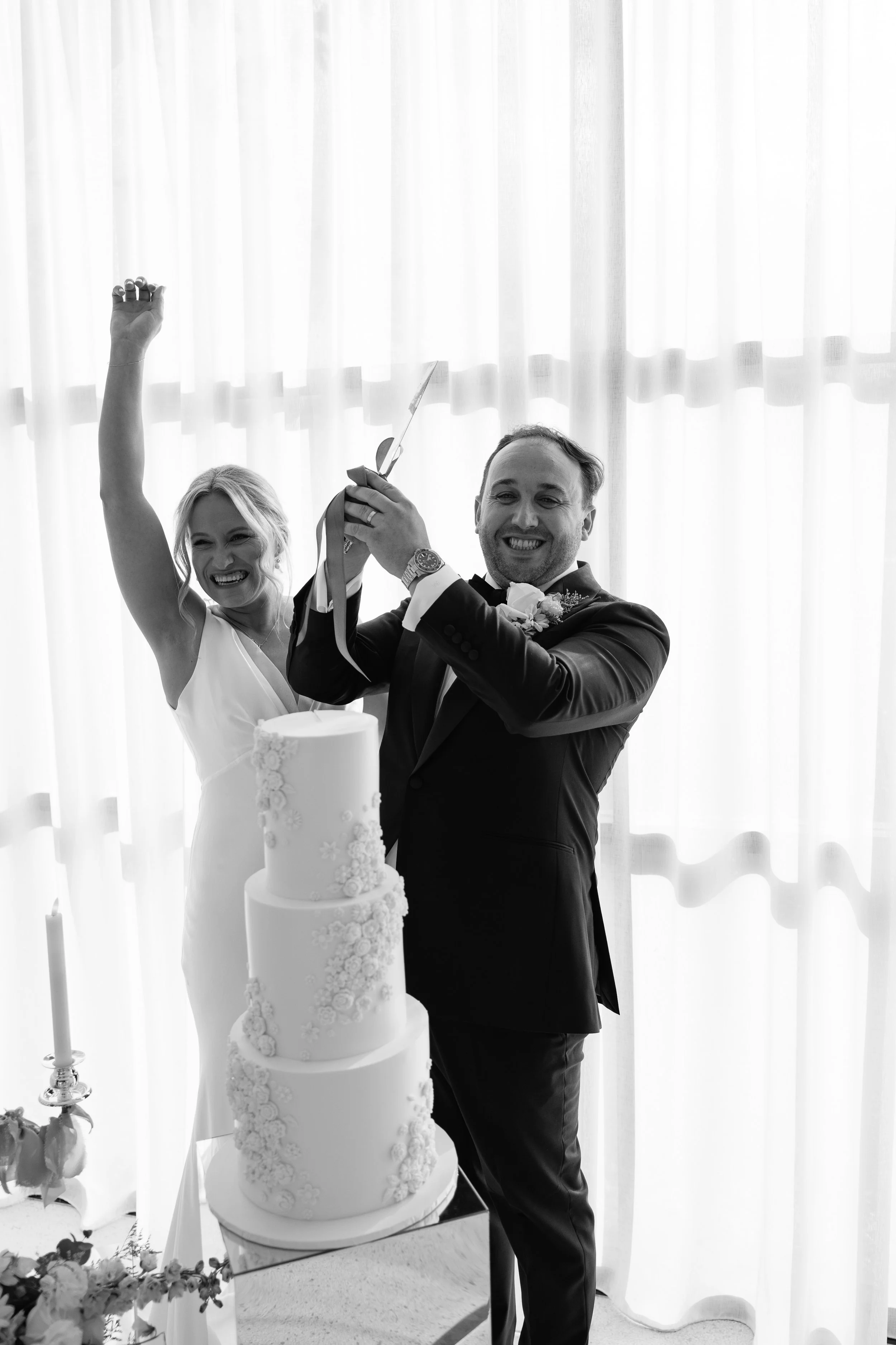 A bride and groom celebrating at their wedding, cutting the wedding cake together in a bright room with large curtains.