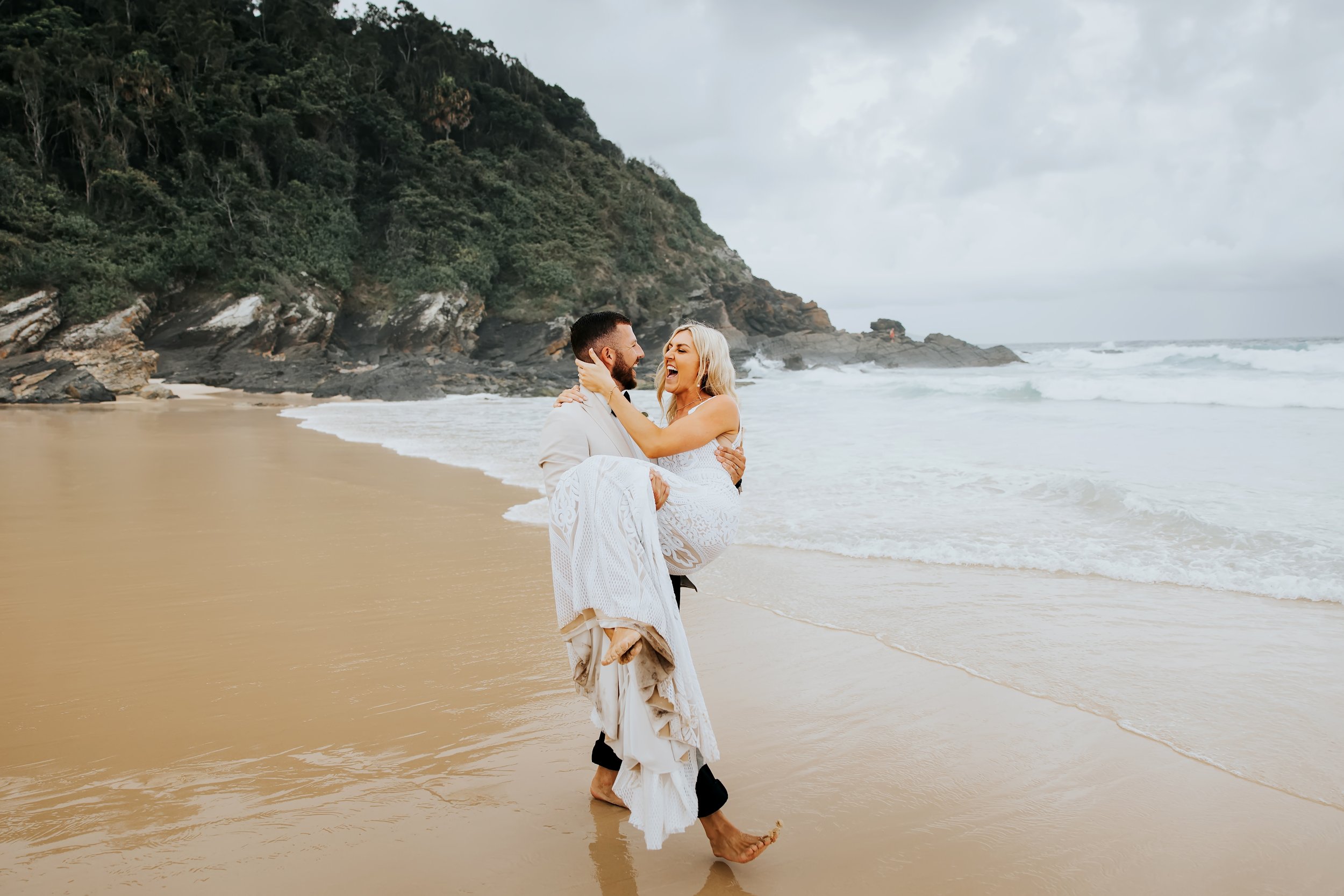 A newlywed couple on a beach, the bride in a white lace dress and the groom in a light-colored suit, smiling and embracing near the ocean with a rocky hillside in the background.