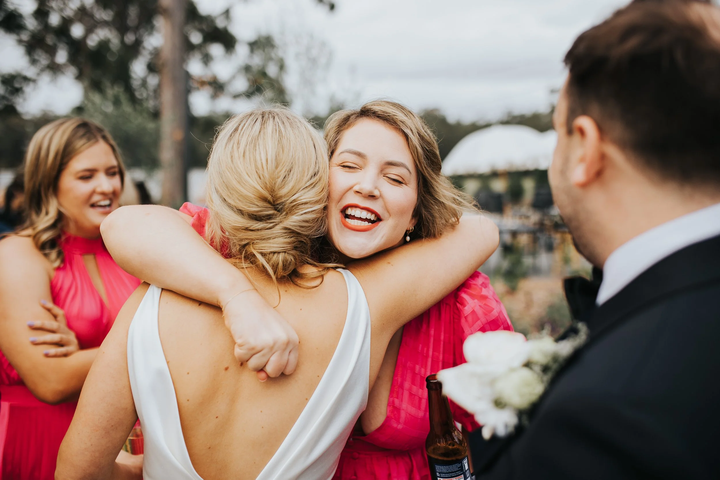Group of people at a celebration, hugging and smiling outdoors, dressed in formal and semi-formal attire.