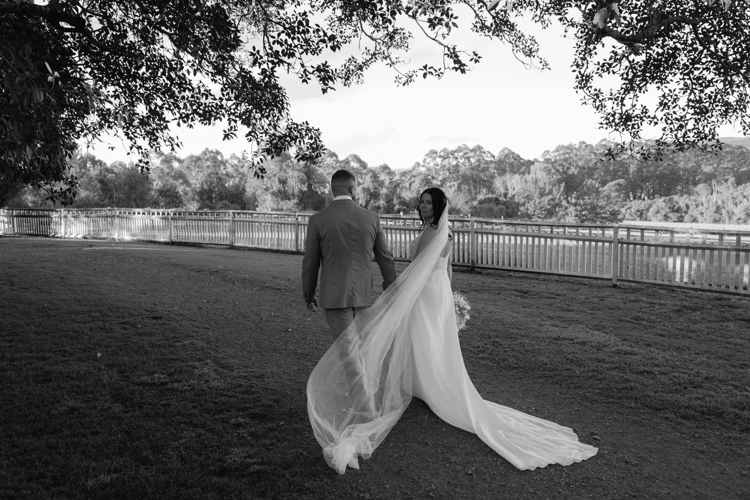 A black and white photo of a bride and groom holding hands outdoors, with a fence and trees in the background, taken during daytime.