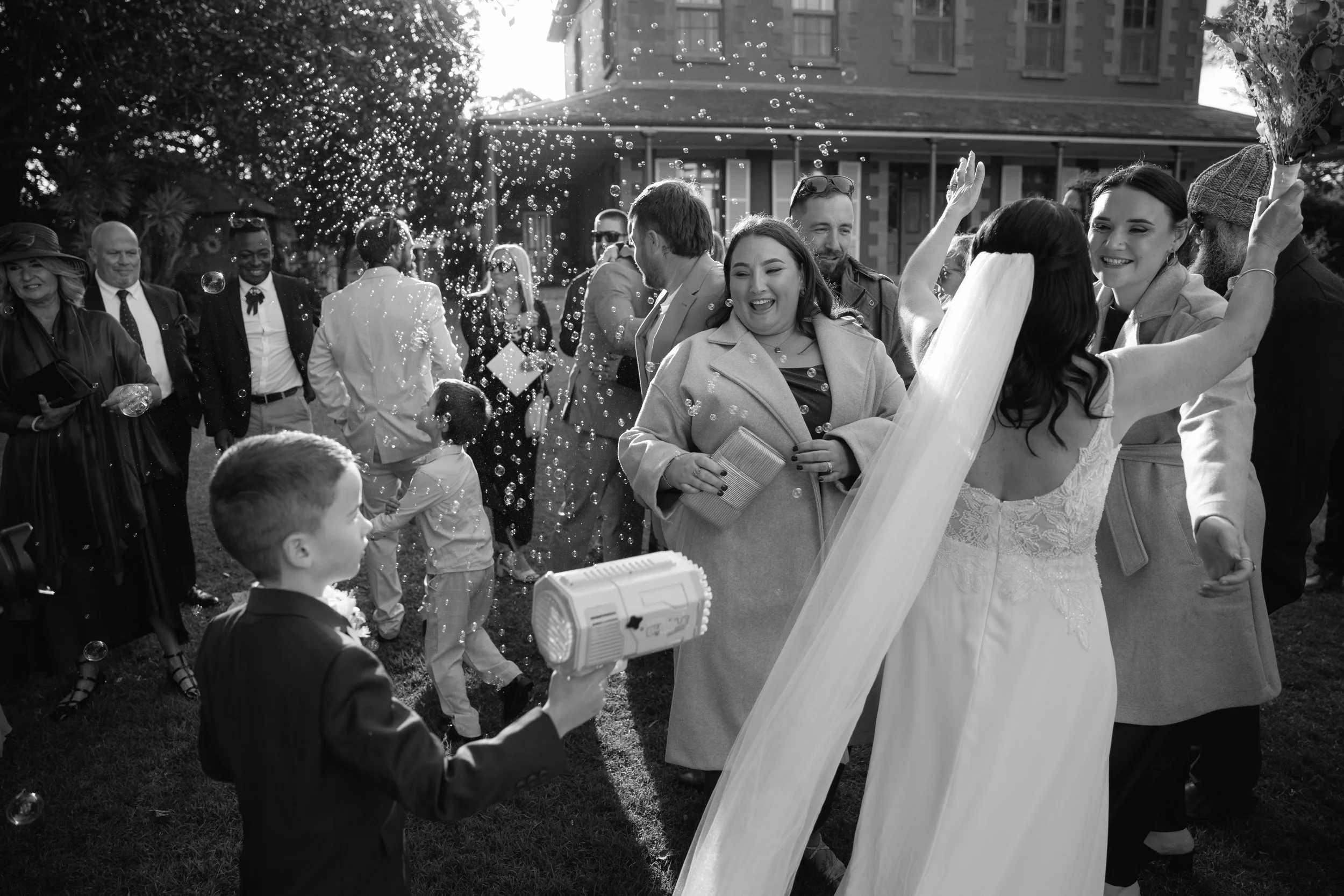 A black and white photo of a wedding celebration outdoors with guests and the bride and groom smiling and interacting.