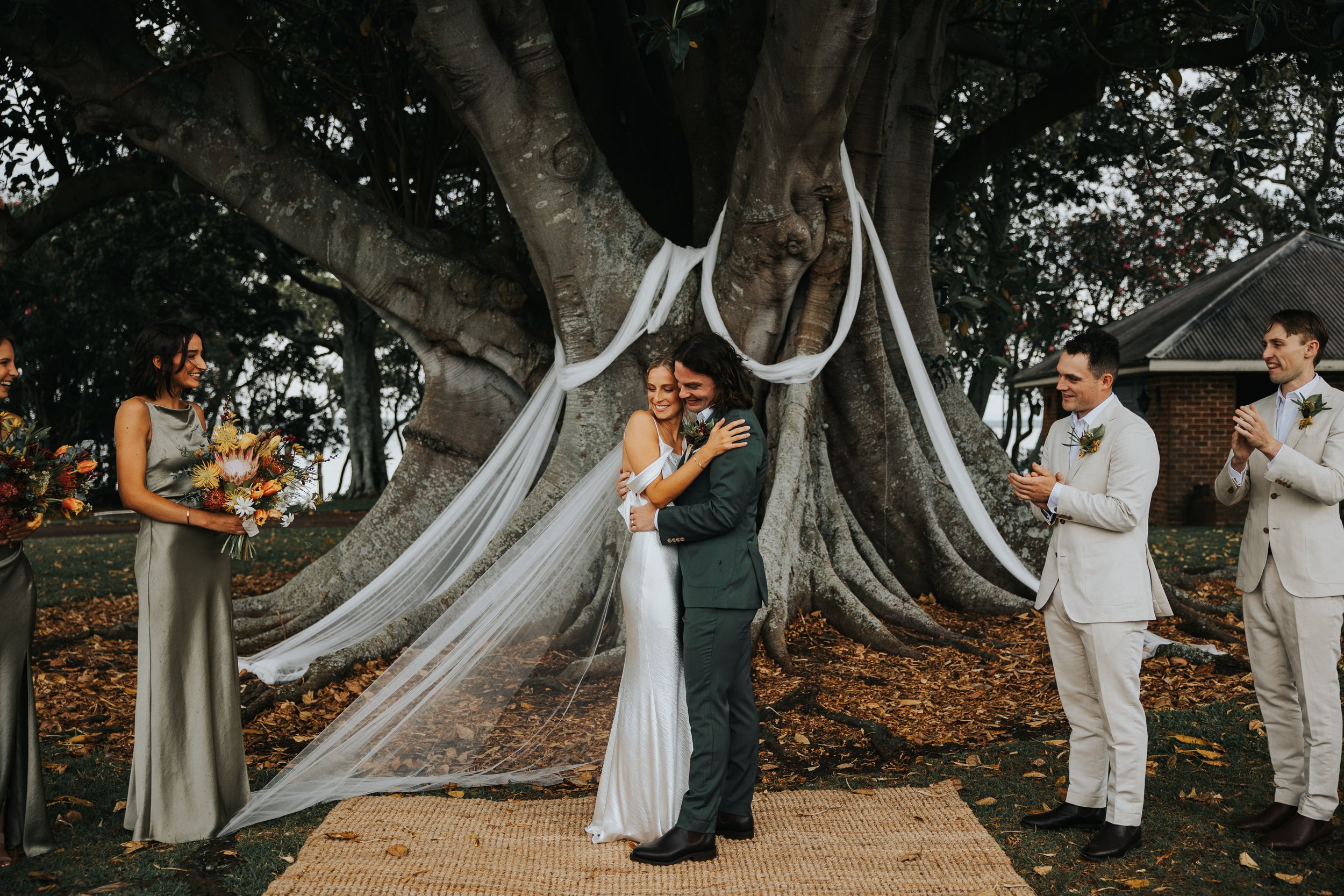 Couple getting married outdoors under a large tree with white fabric decorations. They are hugging, surrounded by wedding party members holding flowers and clapping. The scene includes autumn leaves on the ground and a rustic building in the backgrou