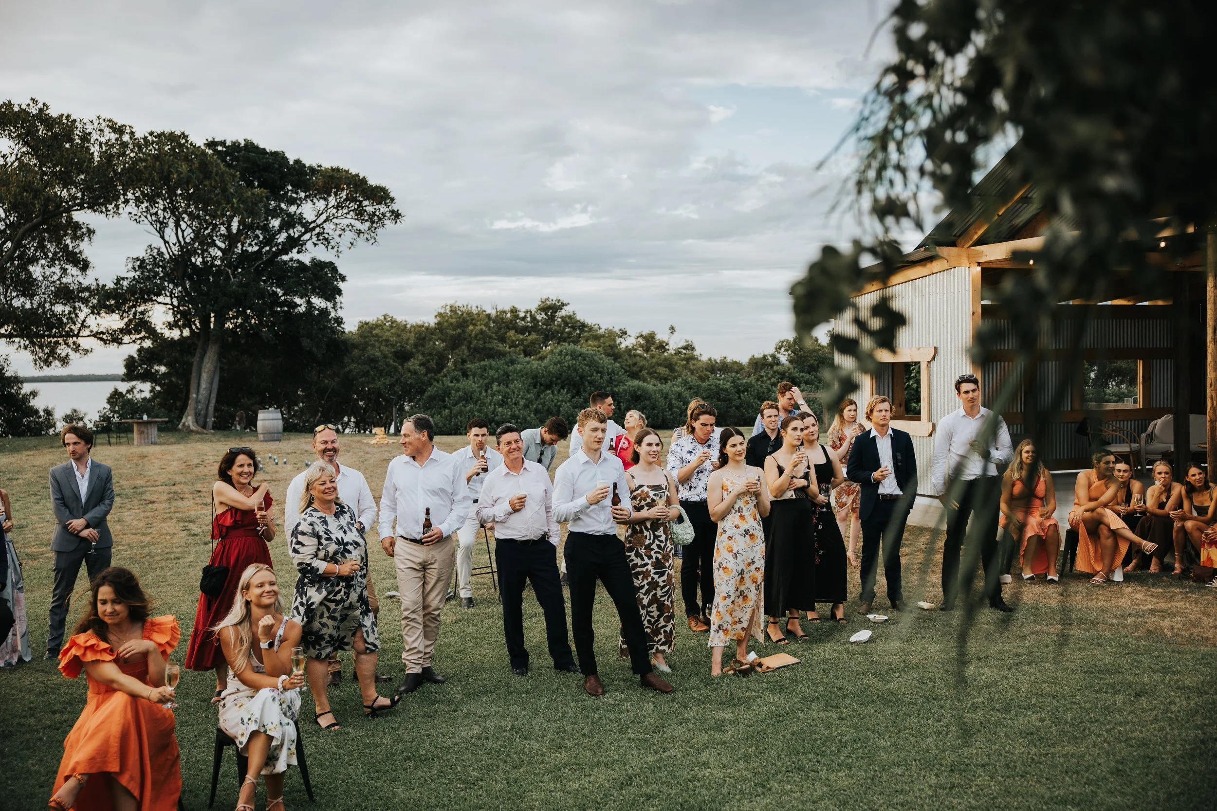 A group of people gathered outdoors for a celebration or event, standing on grass with a large tree and overcast sky in the background. Some people are holding drinks, and there is a rustic building on the right.