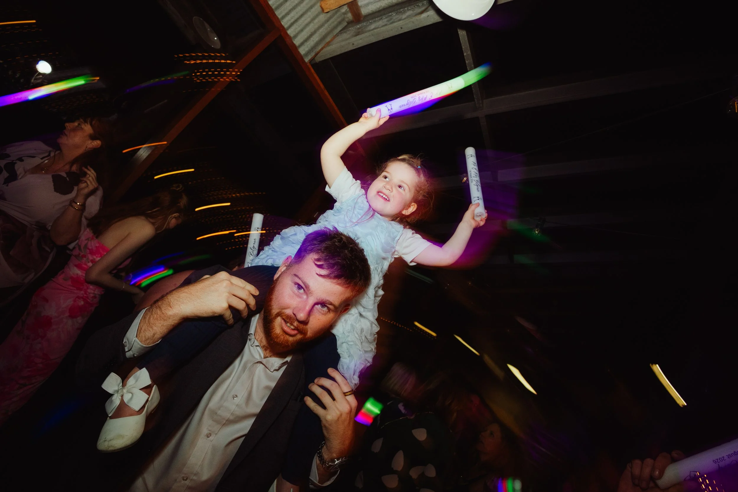 A young girl with a joyful expression holding a glow stick above her head while sitting on a man's shoulders at a dark party or celebration, with colorful lights and other people in the background.