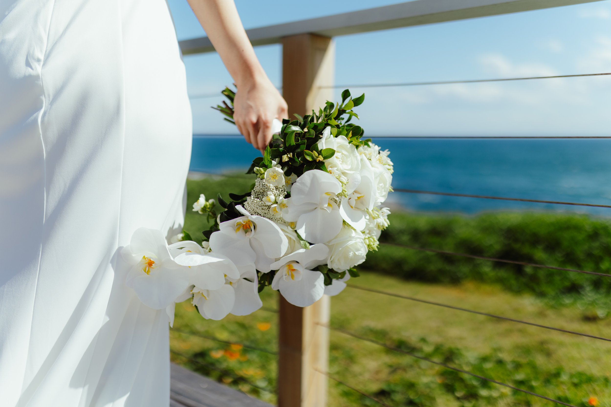 A person wearing a white dress holding a bouquet of white orchids and greenery, standing outdoors on a balcony with ocean and blue sky in the background.