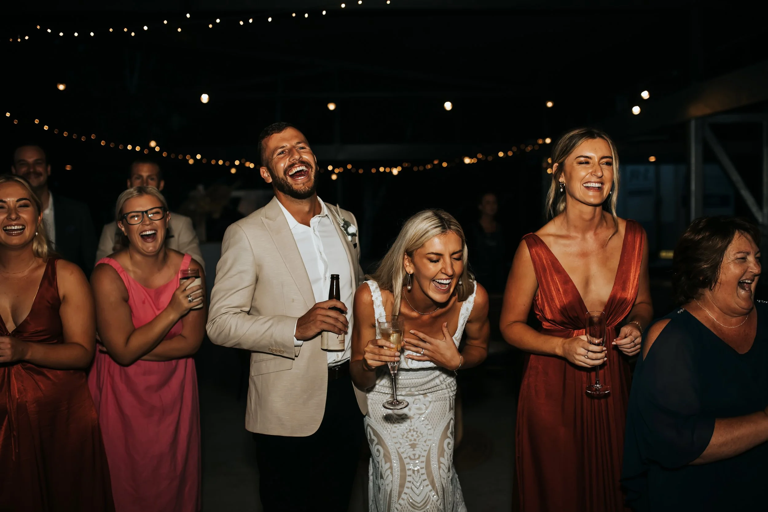 Group of people celebrating at a party, laughing and drinking, with string lights overhead.