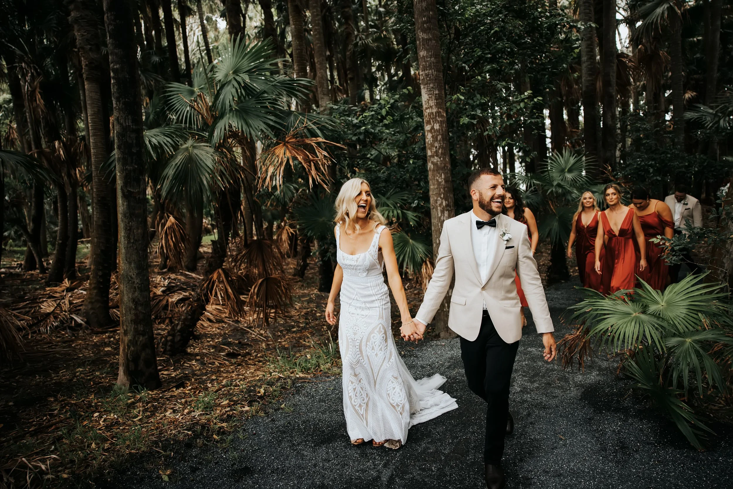 A newly married couple, the bride in a white lace dress and the groom in a white tuxedo with a black bow tie, holding hands and smiling as they walk through a forest with tall trees and lush green foliage. Other bridesmaids in red dresses are followi