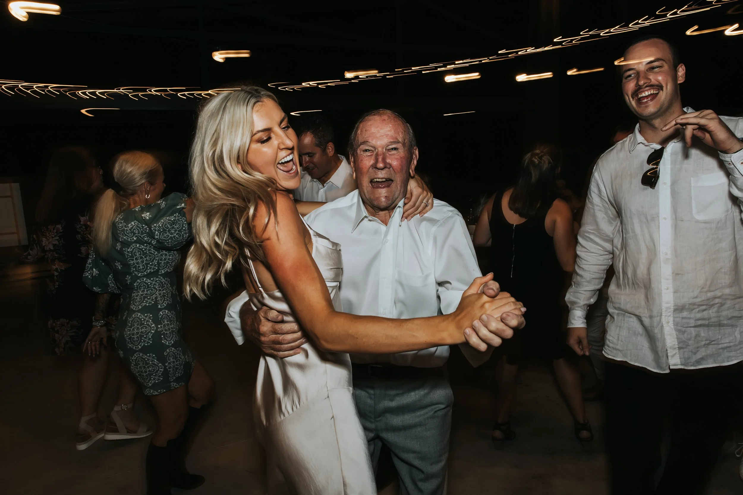 Group of people dancing and celebrating in a dimly lit indoor venue with string lights, including a woman with blonde hair and an older man holding hands and smiling.