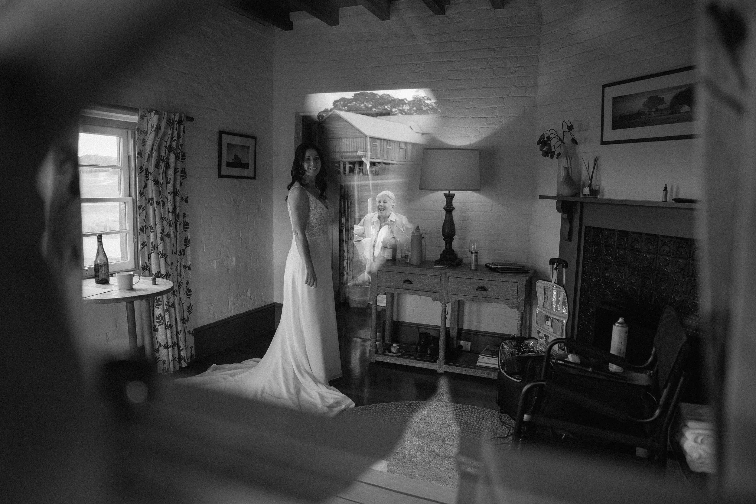 A woman in a wedding dress stands inside a rustic room with a fireplace and framed pictures on the wall, while an elderly woman outside looks through a window and smiles.