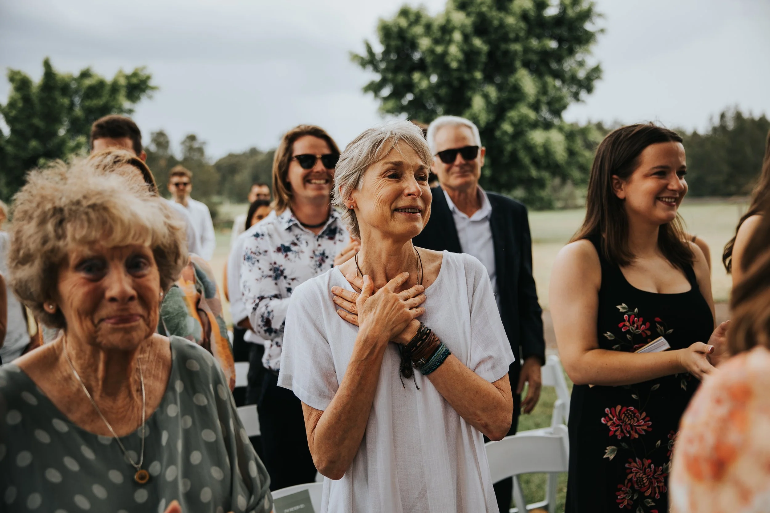 Group of smiling people attending an outdoor event, with a focus on a woman with short gray hair in a white shirt and a woman with dark hair in a black dress, both looking happy and engaged.