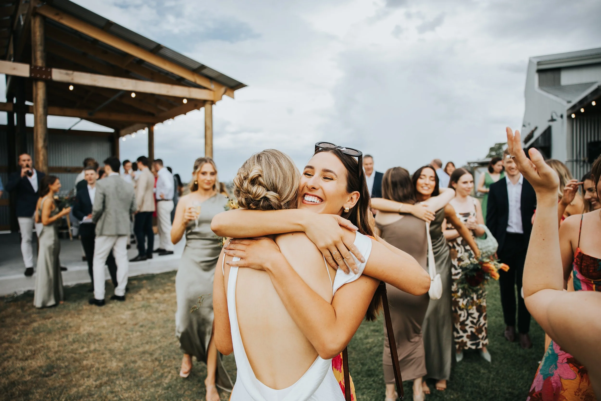 Two women hugging and smiling at a social gathering outdoors with a crowd of people in the background.