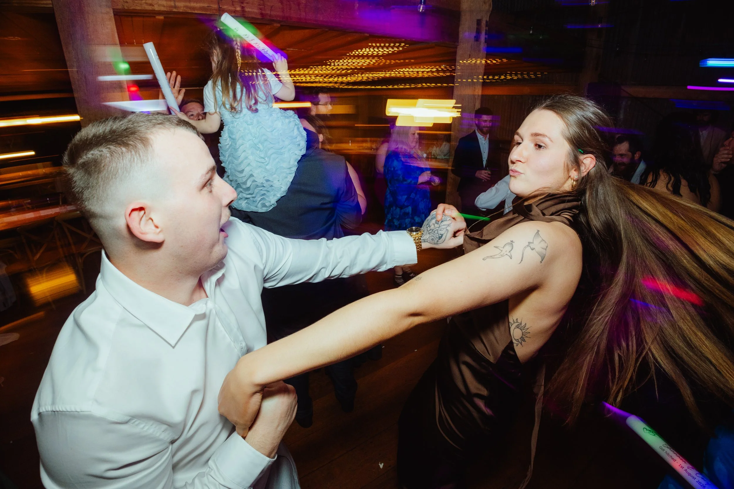 A young man in a white shirt and a woman with tattoos on her arm dance closely with a young girl at a party with colorful lights and other people in the background.