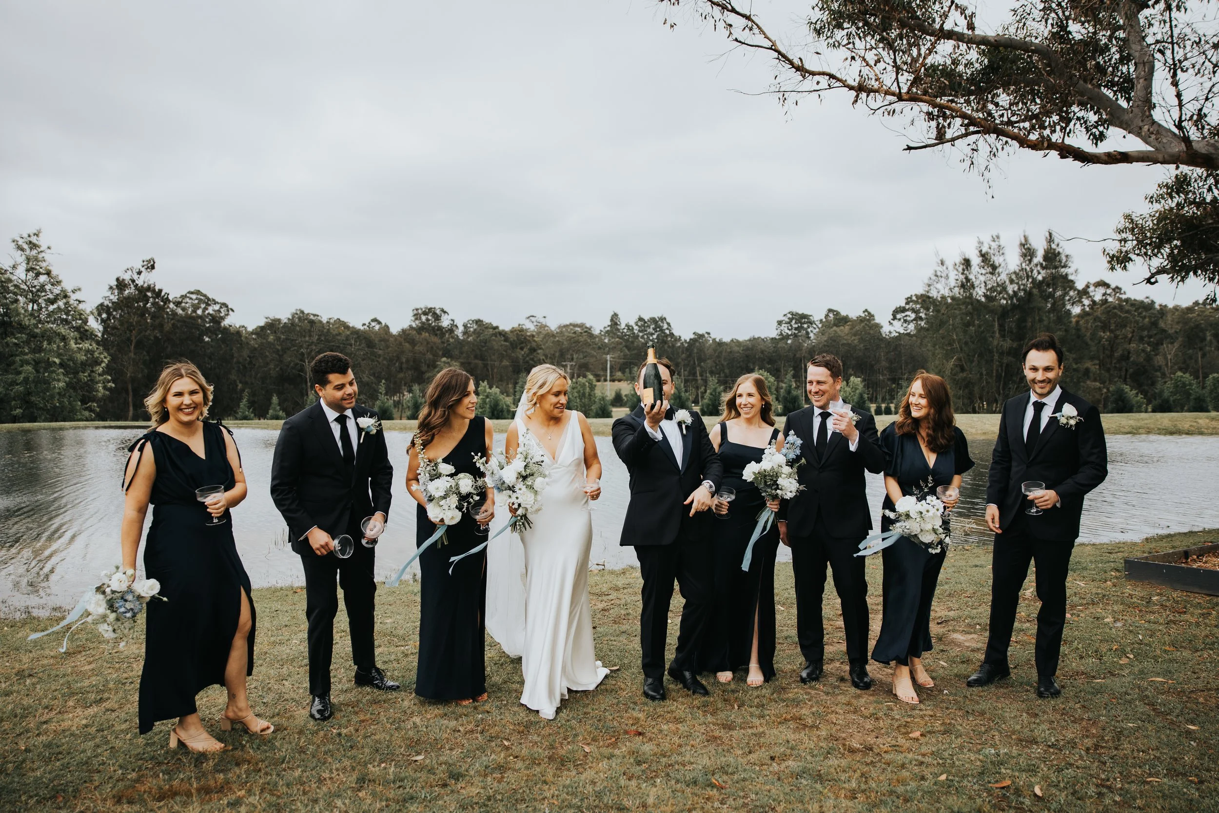 Group of wedding guests walking outdoors by water, dressed in formal attire, some holding glasses, with a tree and cloudy sky in the background.