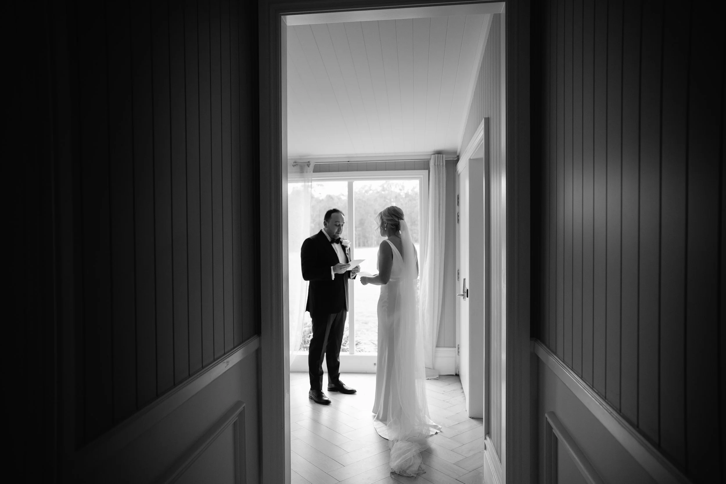 A bride and groom standing together in a doorway, framed by a dark hallway. The groom is reading from a paper, while the bride listens. Natural light illuminates the scene from behind them.