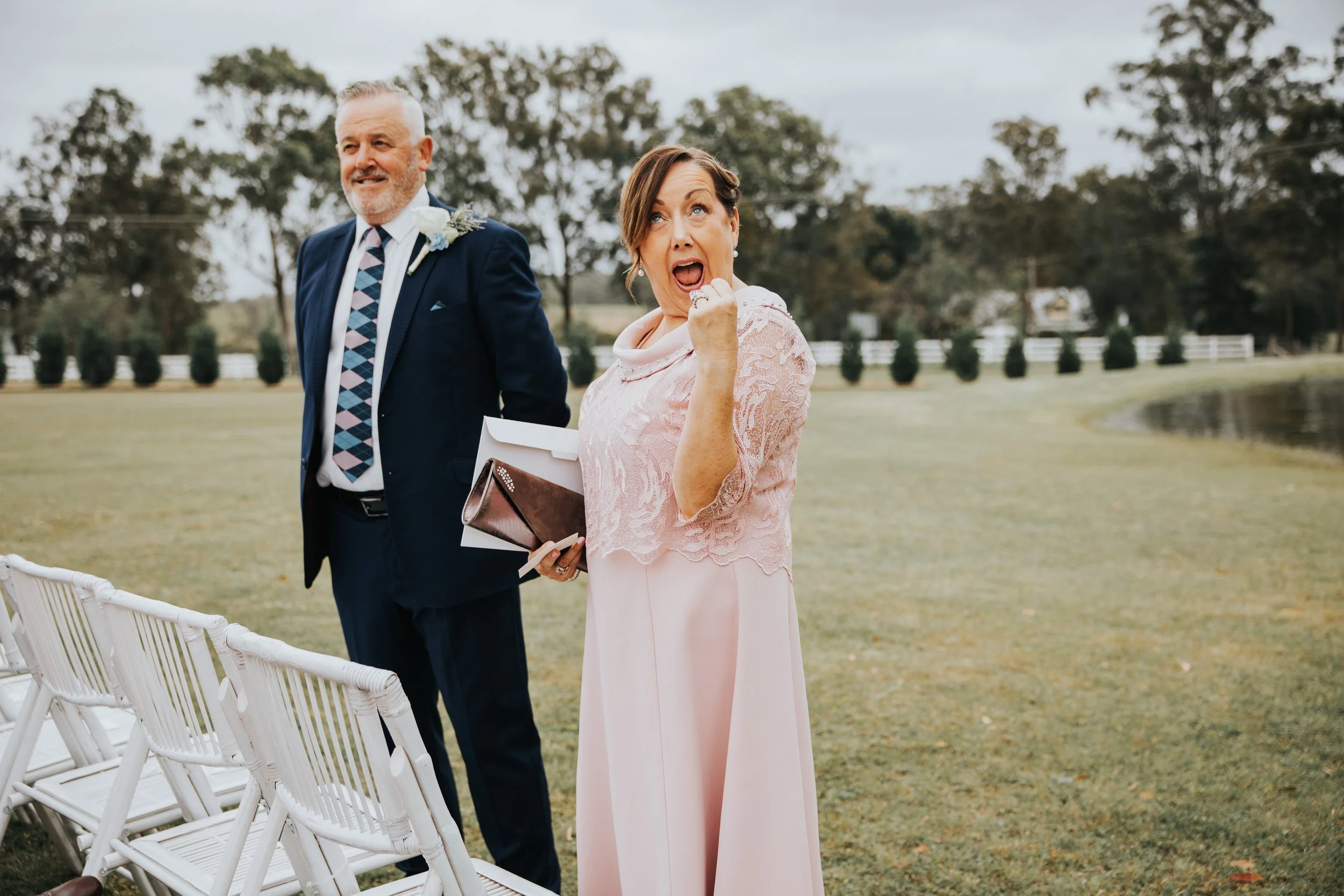 A woman in a pink dress with lace details and a man in a dark suit with a boutonniere on his lapel at an outdoor wedding ceremony. The woman appears surprised and is touching her mouth with her hand. The background features trees, a pond, and a cloud