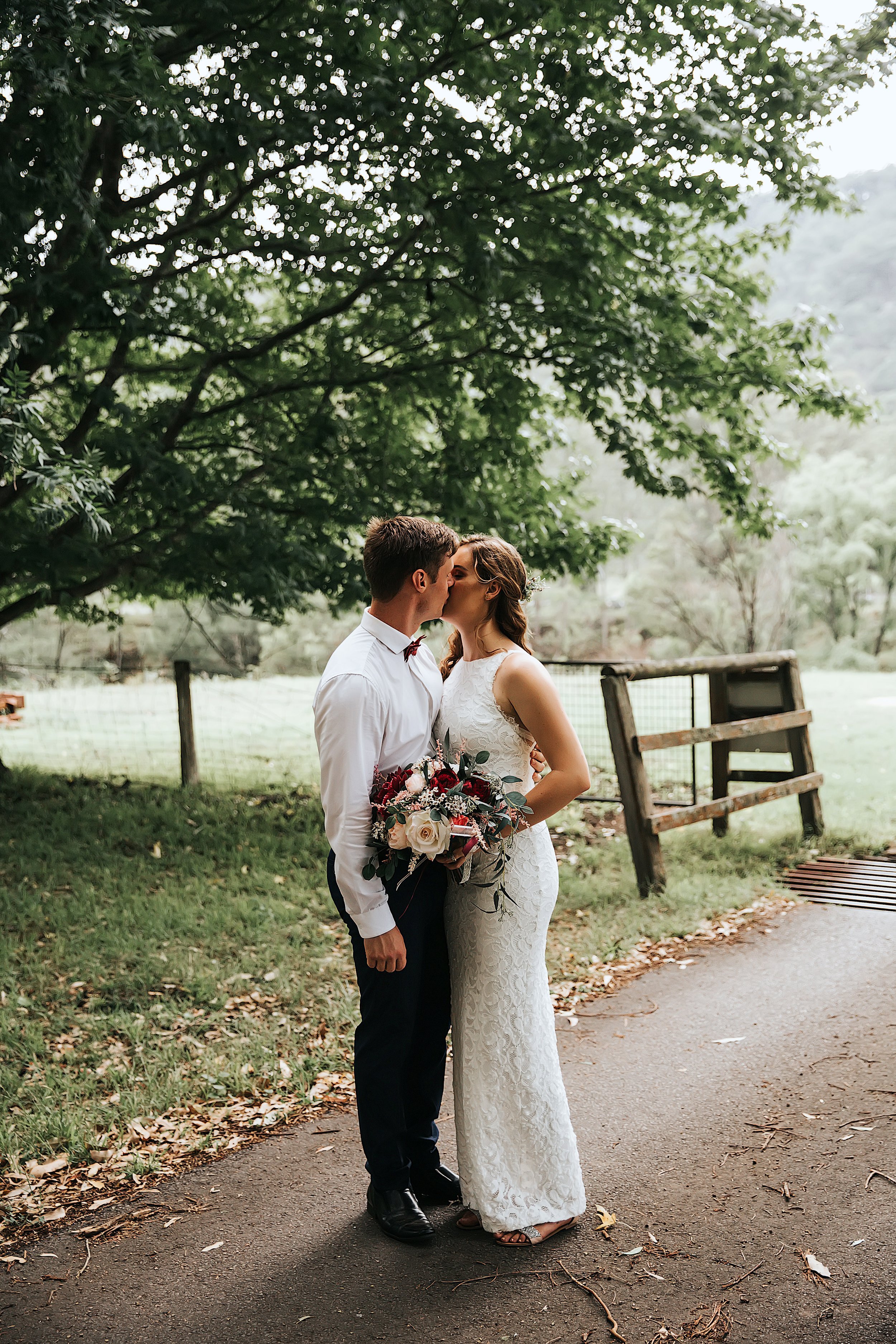 A newlywed couple sharing a kiss outdoors beneath a large leafy tree. The bride is holding a bouquet of flowers and wearing a white lace dress, while the groom is dressed in a white shirt and black pants. They are standing on a paved path with a wood