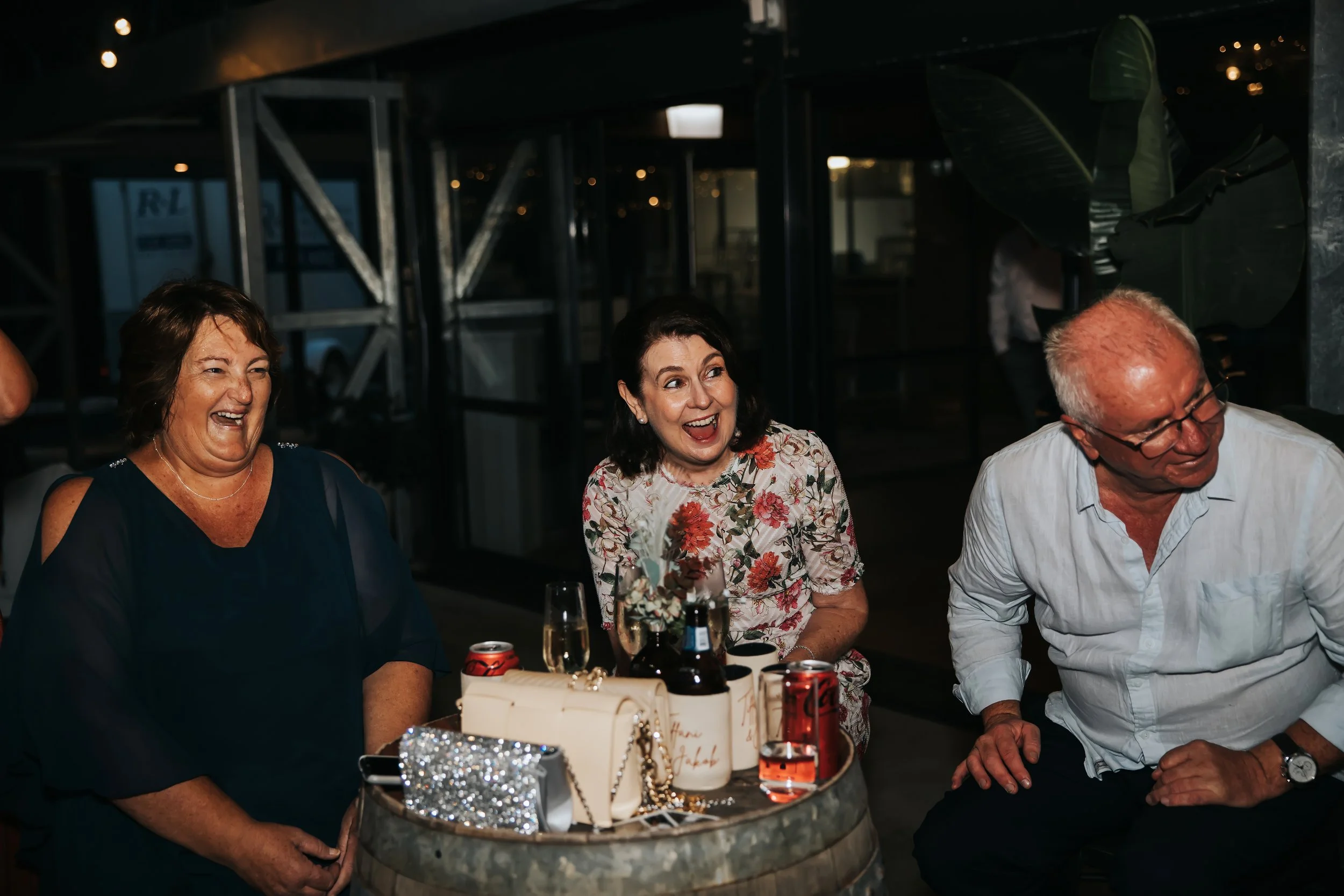 Three smiling adults, two women and one man, are gathered around a table at a celebration or party. The table has various drinks, a small bouquet of flowers, and jewelry as decorations. The background is dimly lit, indicating an indoor setting.