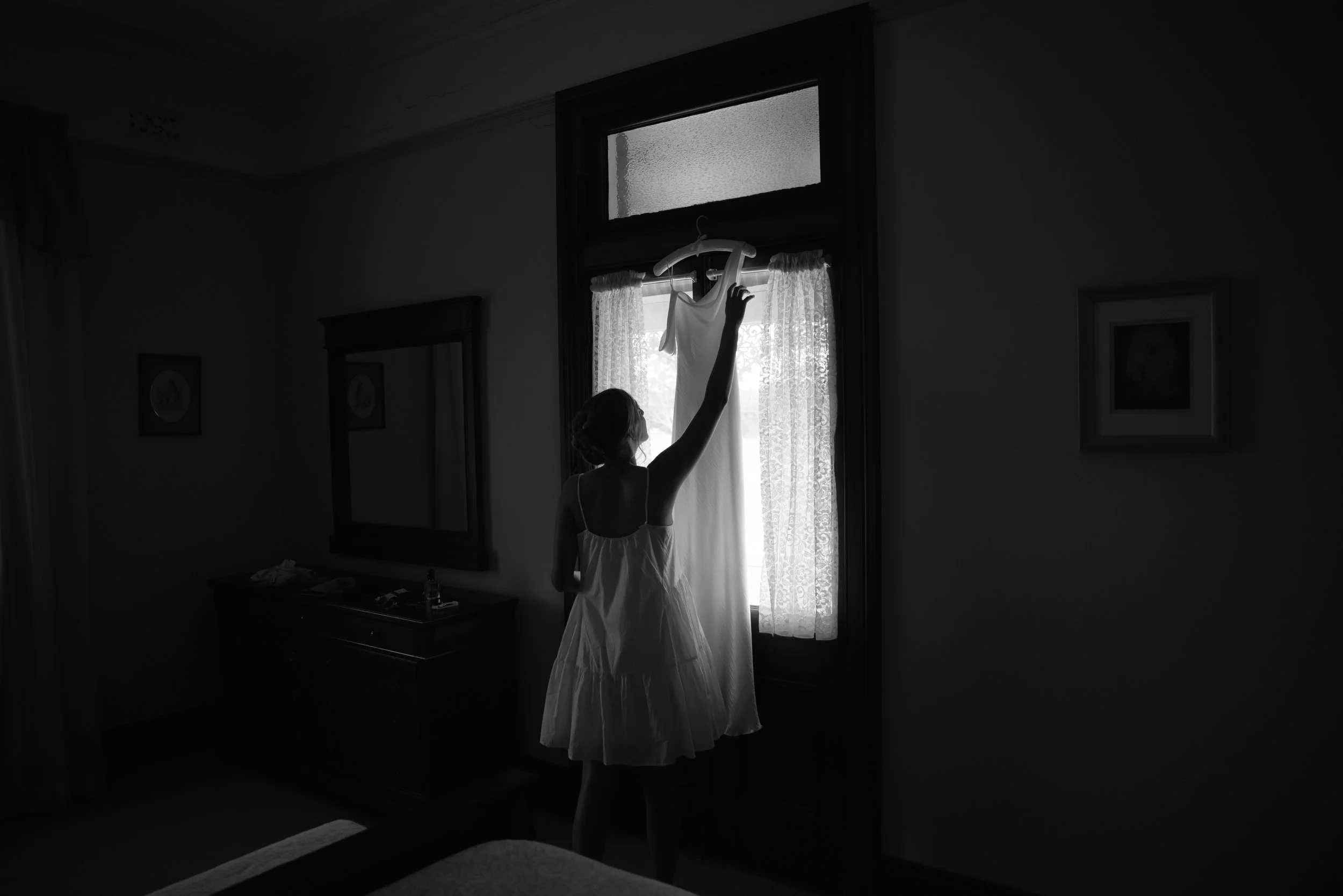 A silhouette of a woman wearing a dress, reaching up to hang a white dress on a hanger near a window with lace curtains.