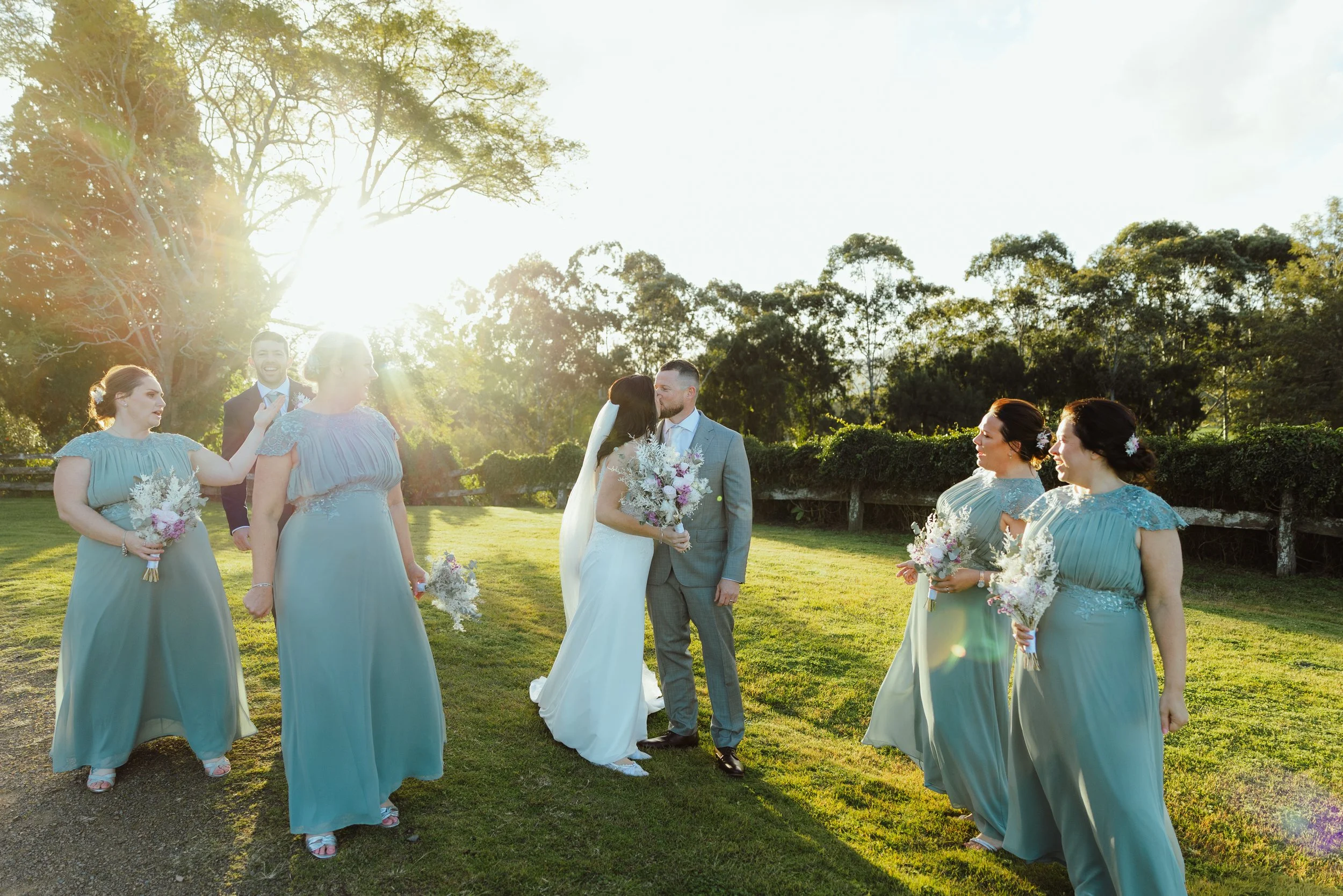 A group of women in matching blue dresses and men in suits, standing outdoors in a grassy area with trees, during a wedding ceremony in the late afternoon.