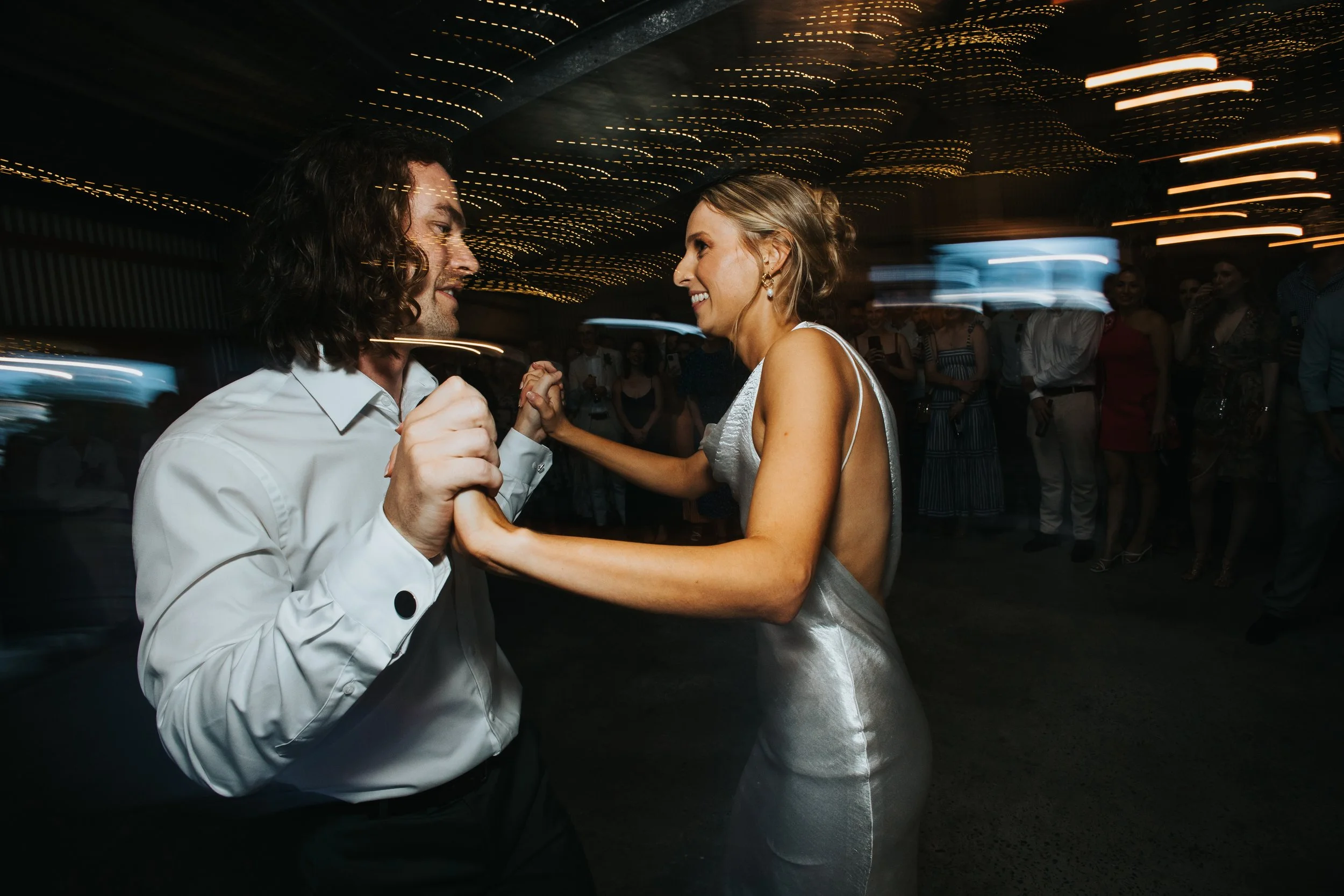 A couple dancing closely at a party, with the man wearing a white shirt and the woman in a silver dress. The background has blurred people and string lights creating a festive atmosphere.
