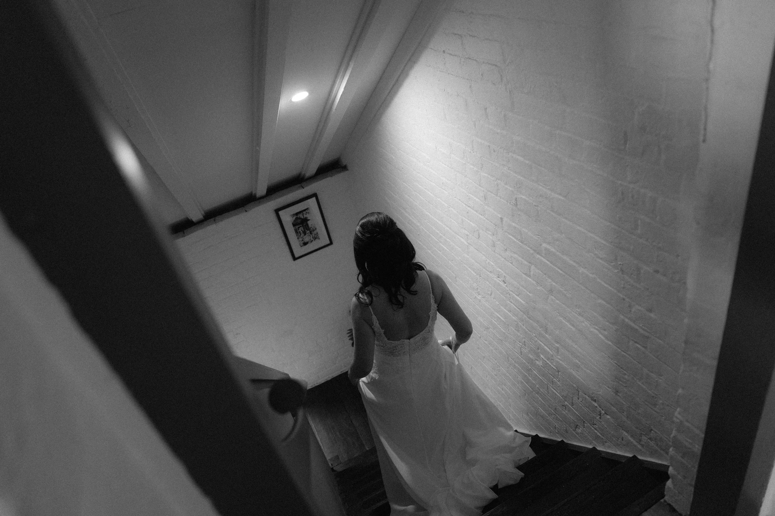 A woman in a wedding dress walking up a staircase with white brick walls, viewed from a camera angle at the bottom of the stairs.