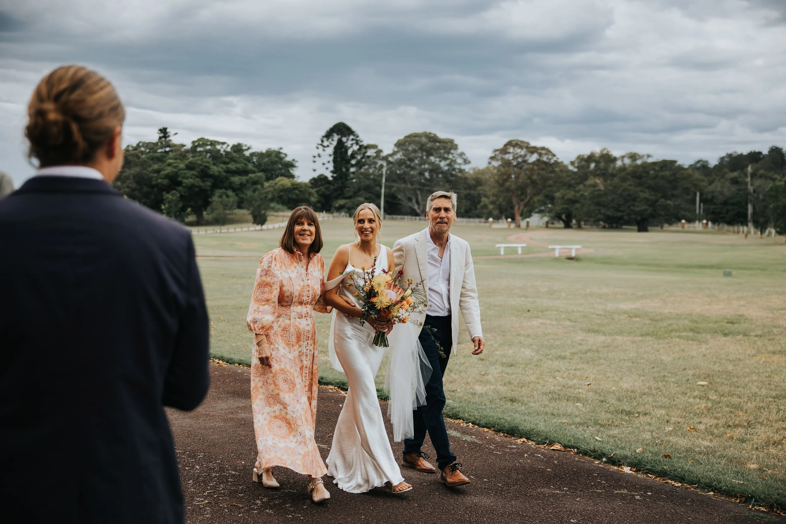 A bride holding a bouquet walking with a man and woman outdoors on a cloudy day, with a man in a suit in the foreground, and trees and open field in the background.