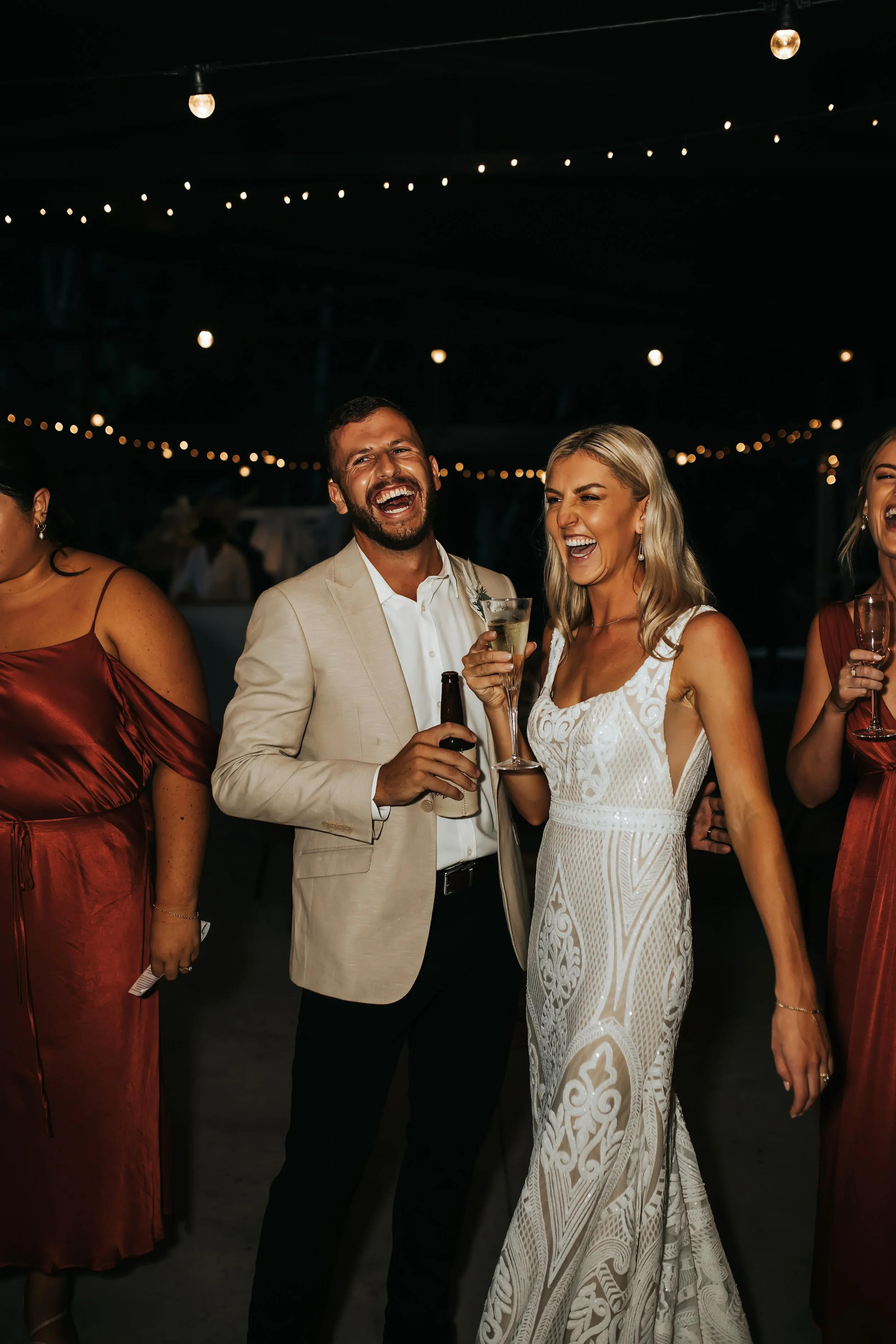 Group of people celebrating at an event under string lights, smiling, holding drinks, dressed in formal attire.