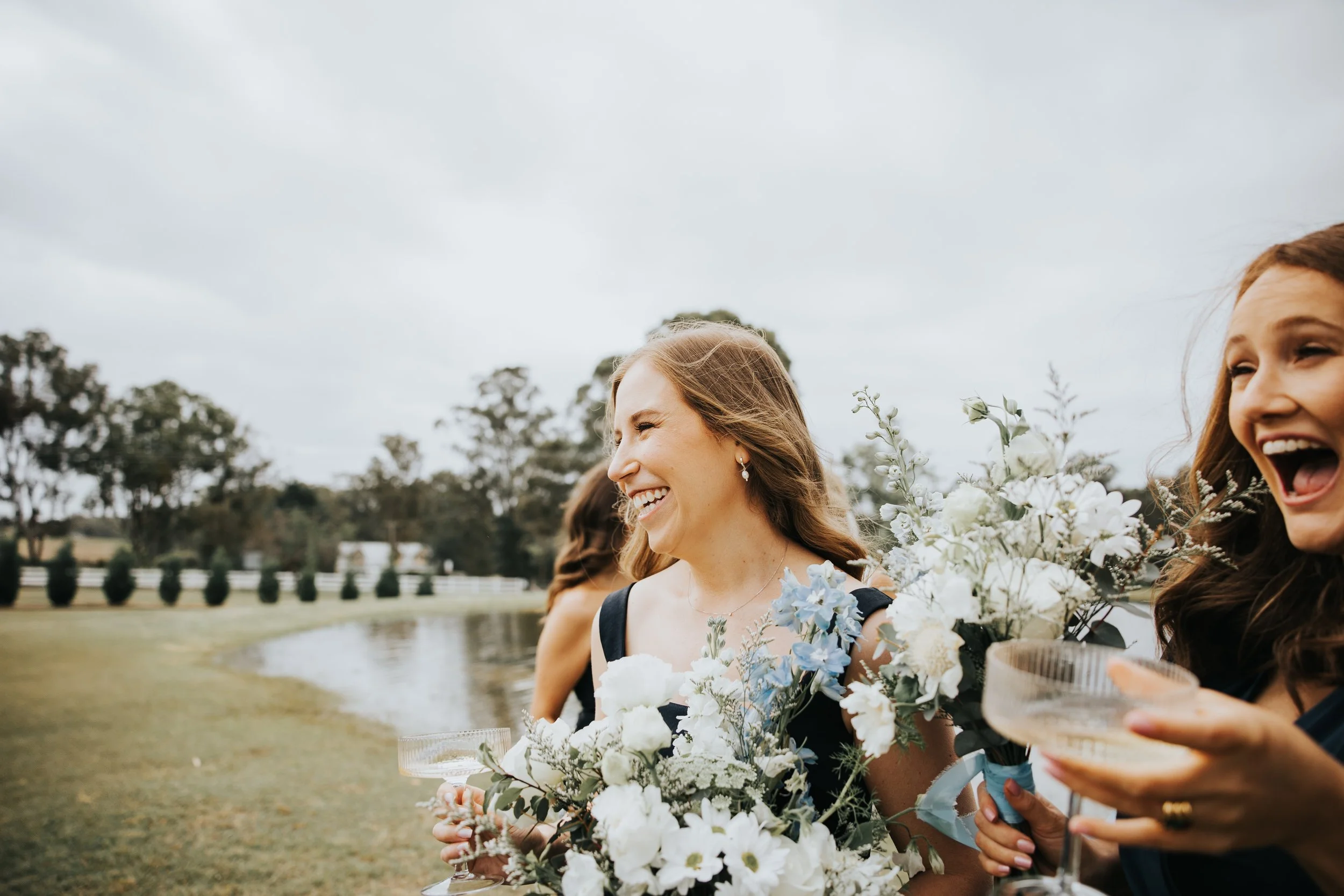 Women in formal dresses holding flowers and glasses, smiling outdoors near a body of water and trees under a cloudy sky.