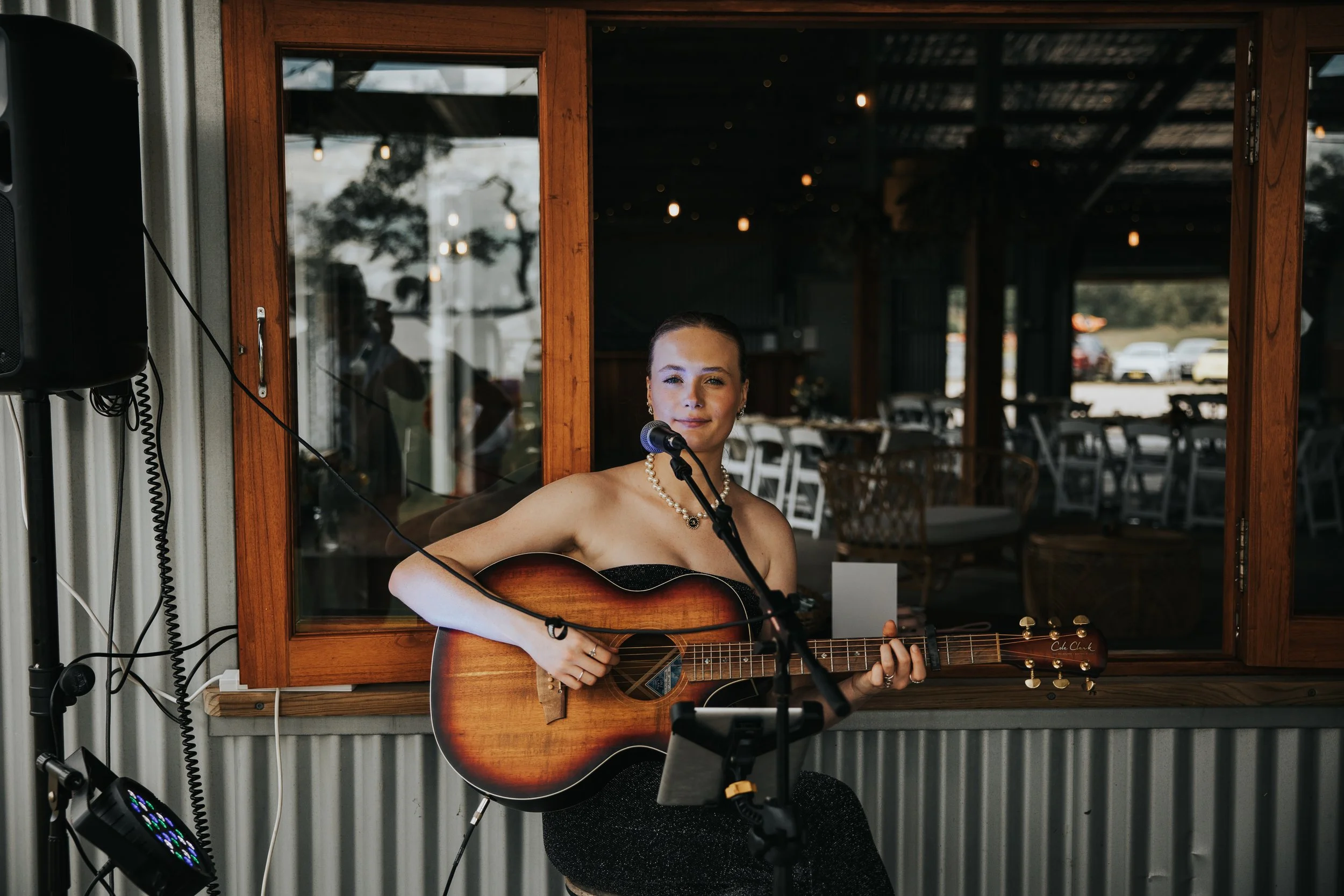 A woman wearing a black dress and pearl necklace playing an acoustic guitar, standing inside a venue with wooden framed windows and hanging lights outside.