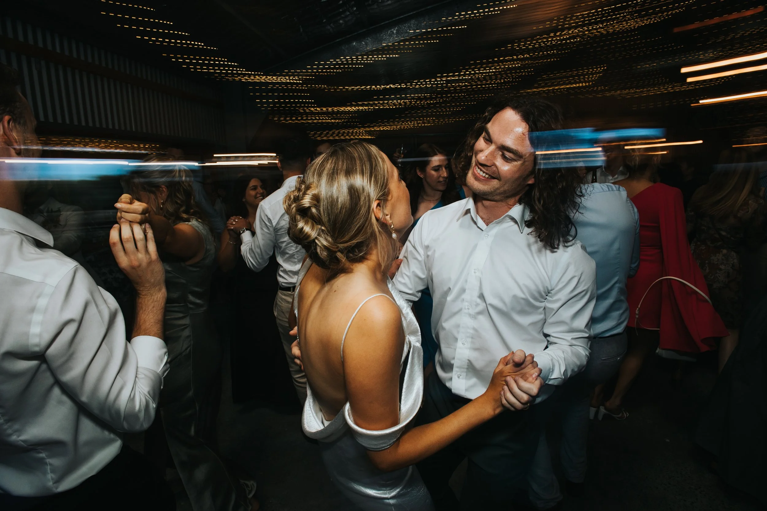 A couple dancing together at a lively party with dim lighting and a ceiling decorated with string lights, surrounded by other people enjoying the celebration.