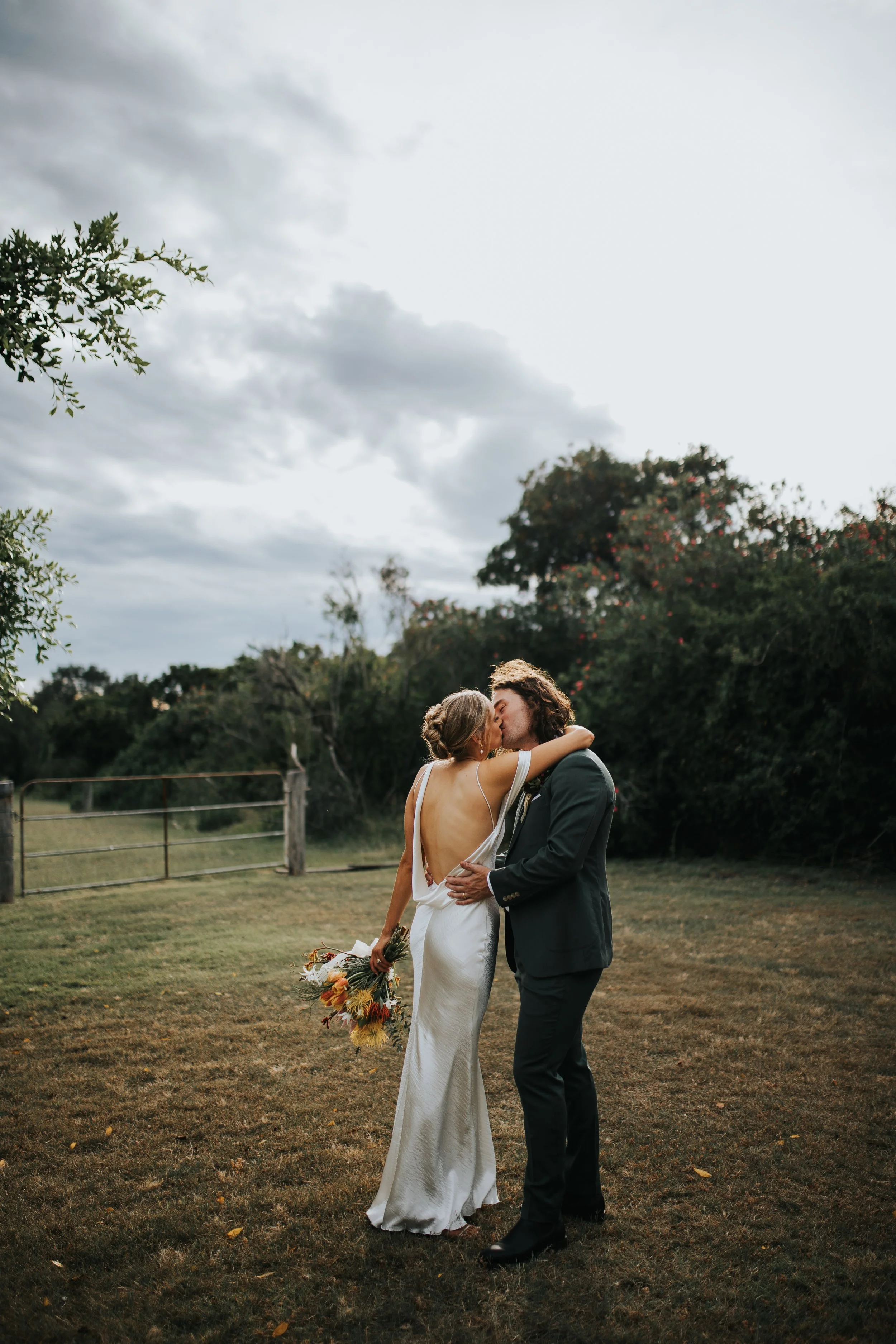 A bride and groom share a kiss outdoors on a grassy field, with trees and a cloudy sky in the background during daytime. The bride holds a bouquet of flowers and wears a white gown, while the groom wears a dark suit.