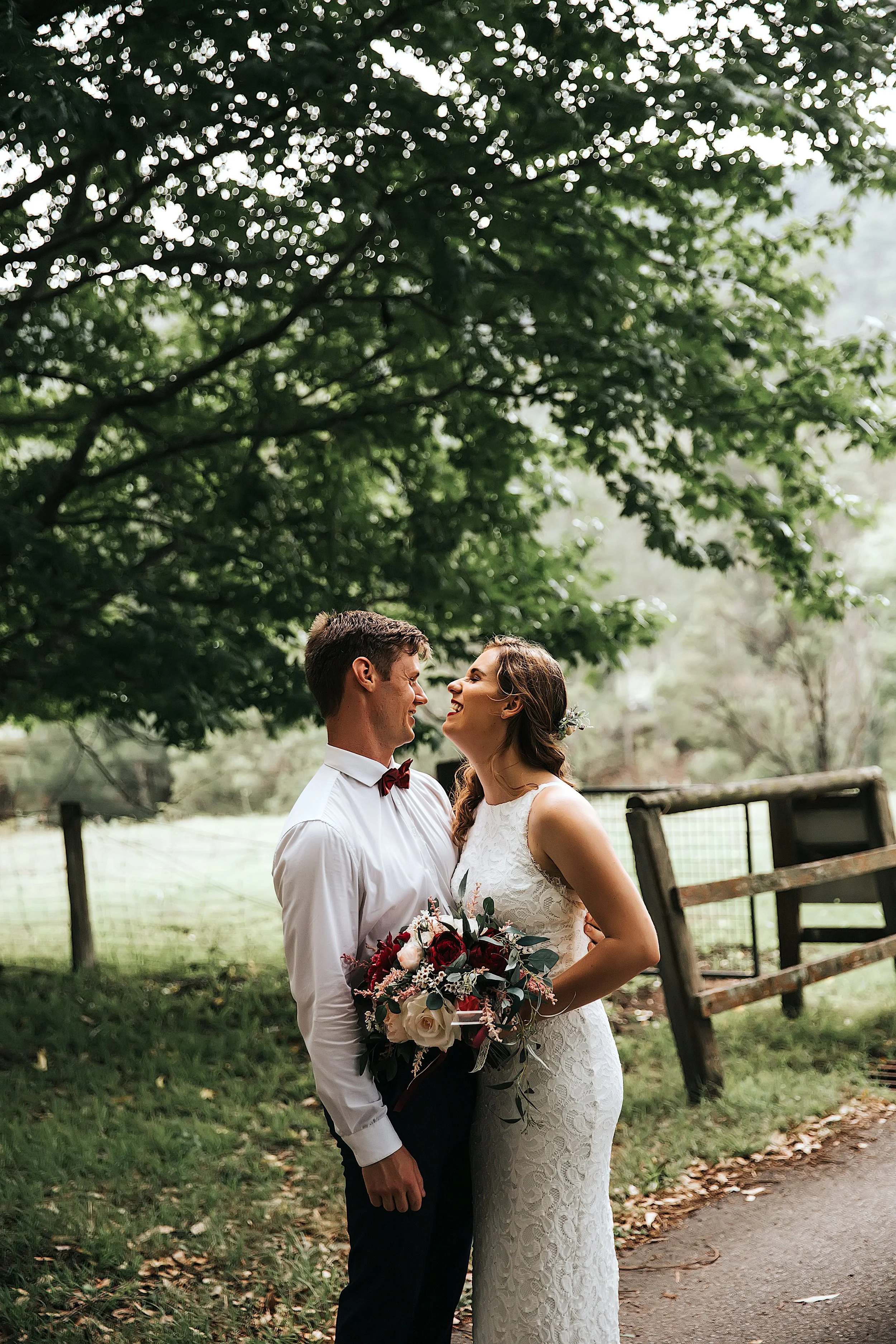 A bride and groom are standing close together outdoors, smiling and looking into each other's eyes, surrounded by greenery with trees overhead and a rustic wooden fence in the background. The bride is holding a bouquet of flowers.