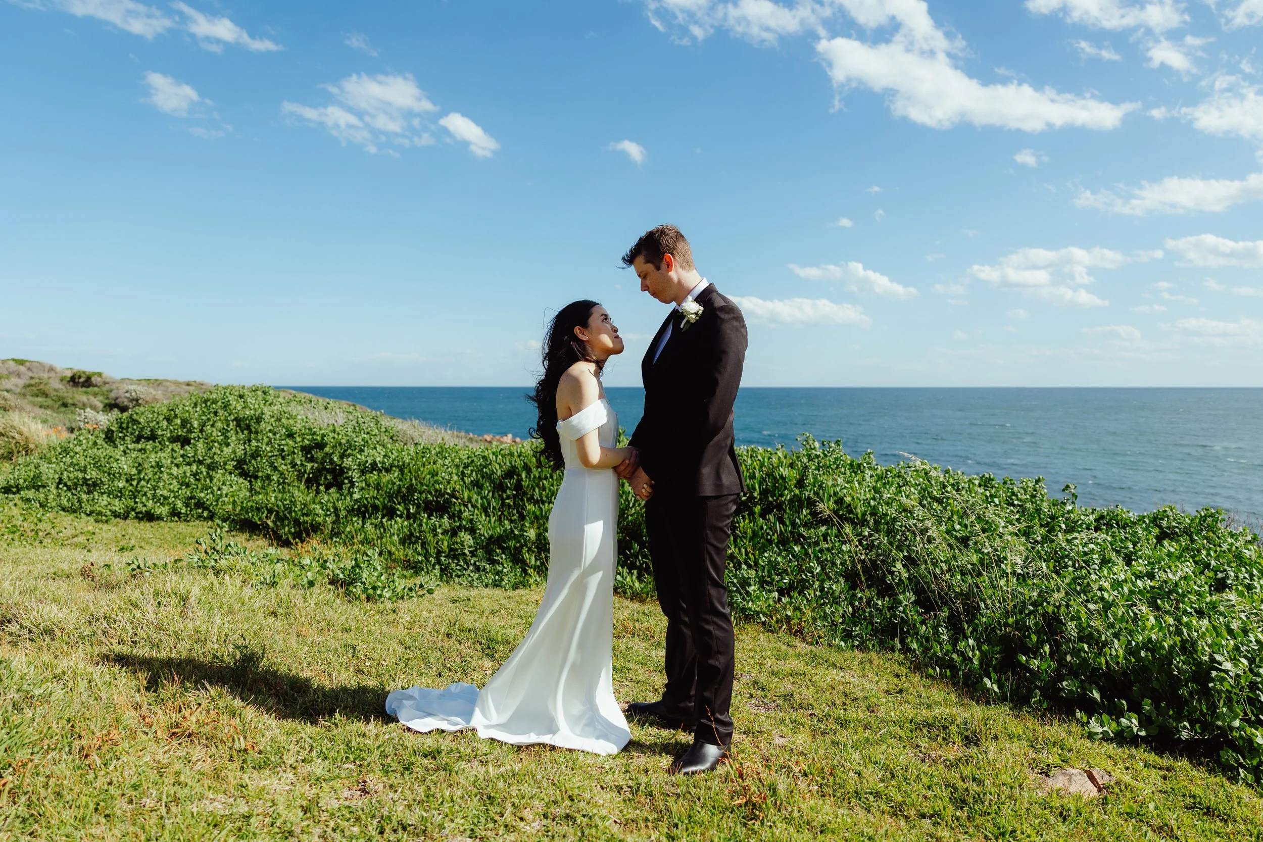 A bride and groom standing outdoors on a grassy area near the ocean, holding hands and gazing at each other, with a blue sky and scattered clouds overhead.