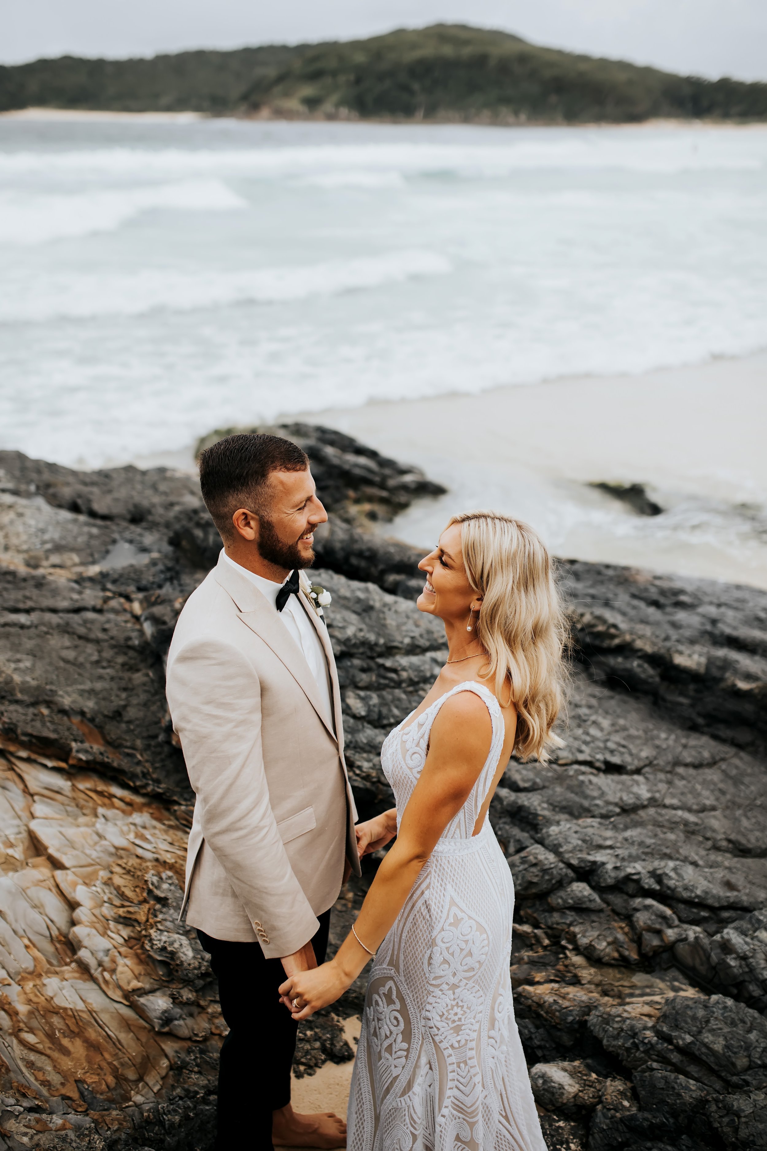 A couple dressed in wedding attire standing hand in hand on a rocky beach, smiling and gazing at each other, with the ocean and a hill in the background.